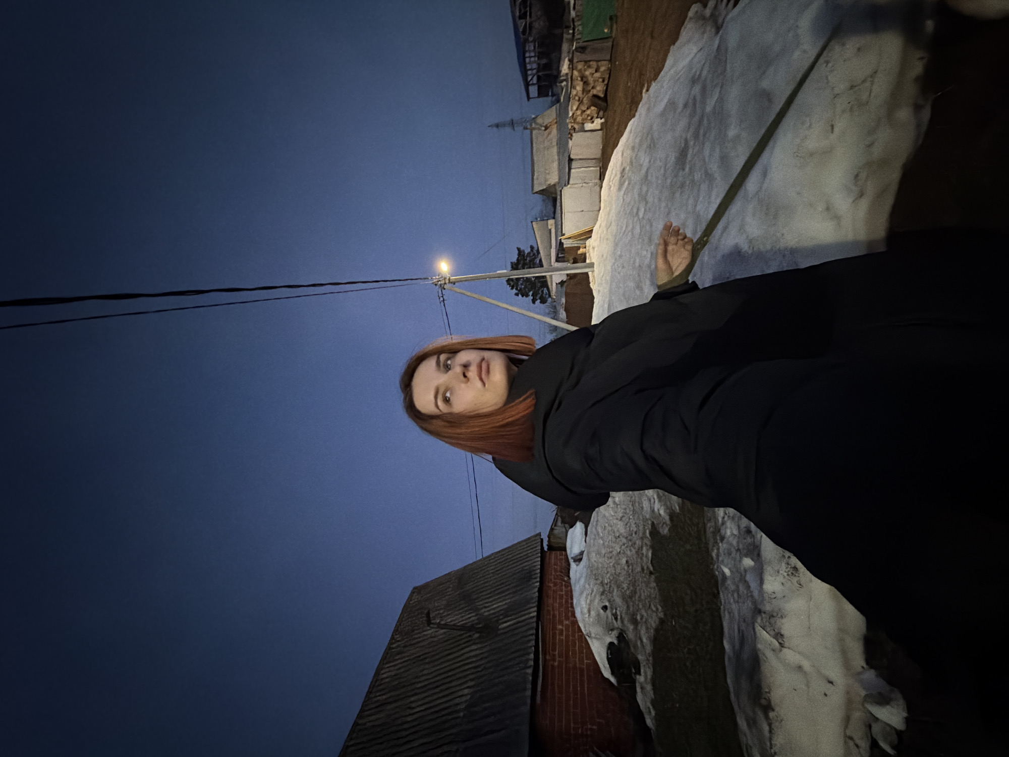 A woman with red hair stands outdoors at dusk in Chaikovsky, Russia. She wears a black jacket. Her bare foot rests on a snow-covered surface. A utility pole with a lit streetlight is visible in the background. There are buildings and stacked wood visible behind the snow. Overhead, power lines extend across the scene. A building with a dark, corrugated roof is in the lower left.