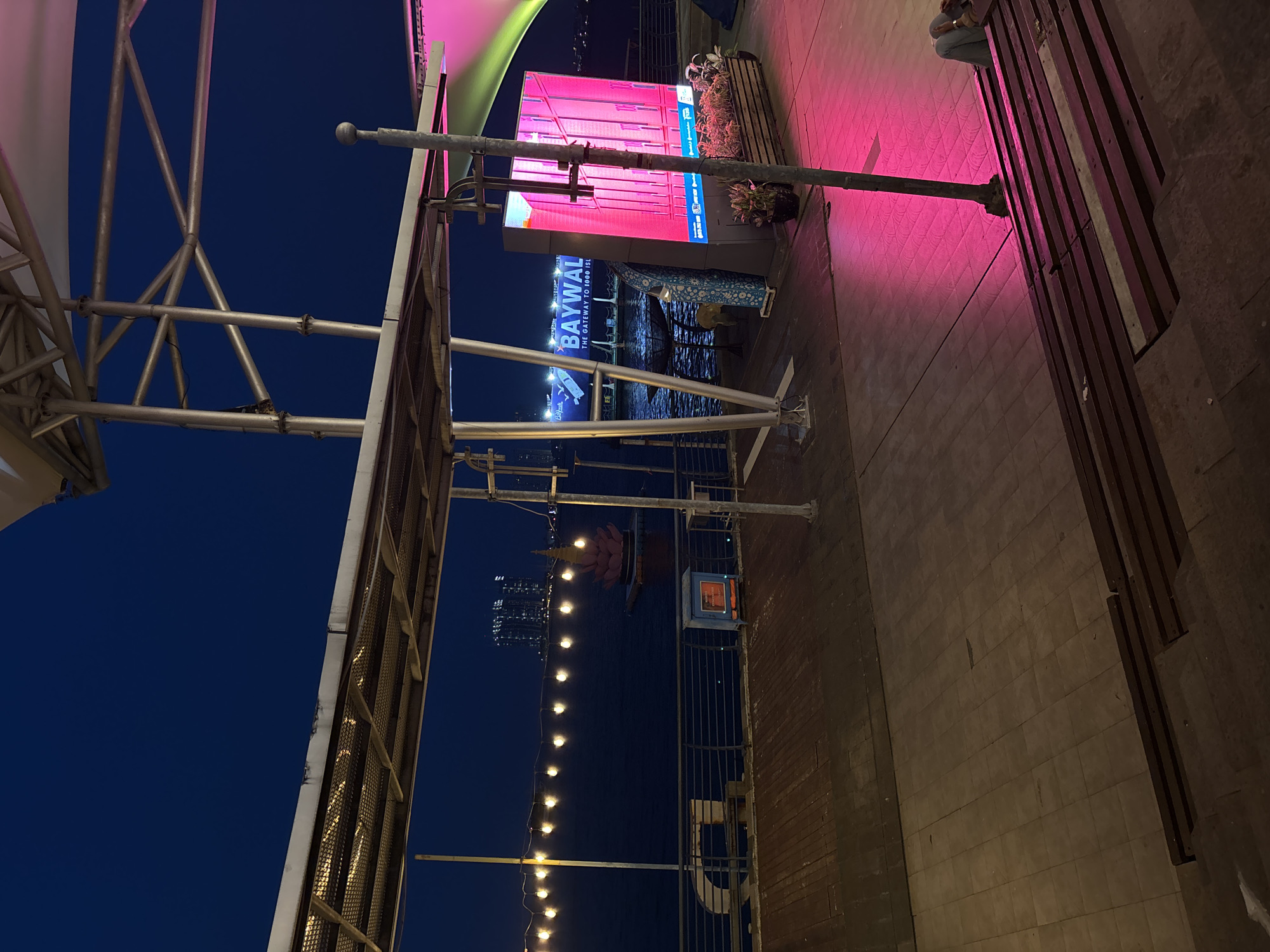 The image shows a night scene at Baywalk Mall in North Jakarta, Indonesia. The foreground features structural elements of a building or covering, including metal beams and grating against a dark blue sky. A walkway is visible, illuminated by pink and white lighting, alongside a sign that says "BAYWALK, THE GATEWAY TO 1000 ISLANDS". String lights stretch into the distance over the water. A bench sits on the walkway, and a person is seated at the end of the bench. Buildings are visible in the background.