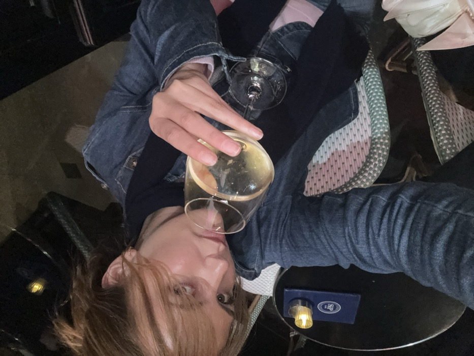 A woman holds a glass of white wine close to her lips, partially tilted, with a second, empty glass resting on her lap. She wears a denim jacket over a pink top, and a dark garment, likely jeans, on her lower body. She sits in a chair with a patterned seat. A small round table to her right holds a blue book and a lamp. The image appears to be a selfie taken at an unusual angle.
