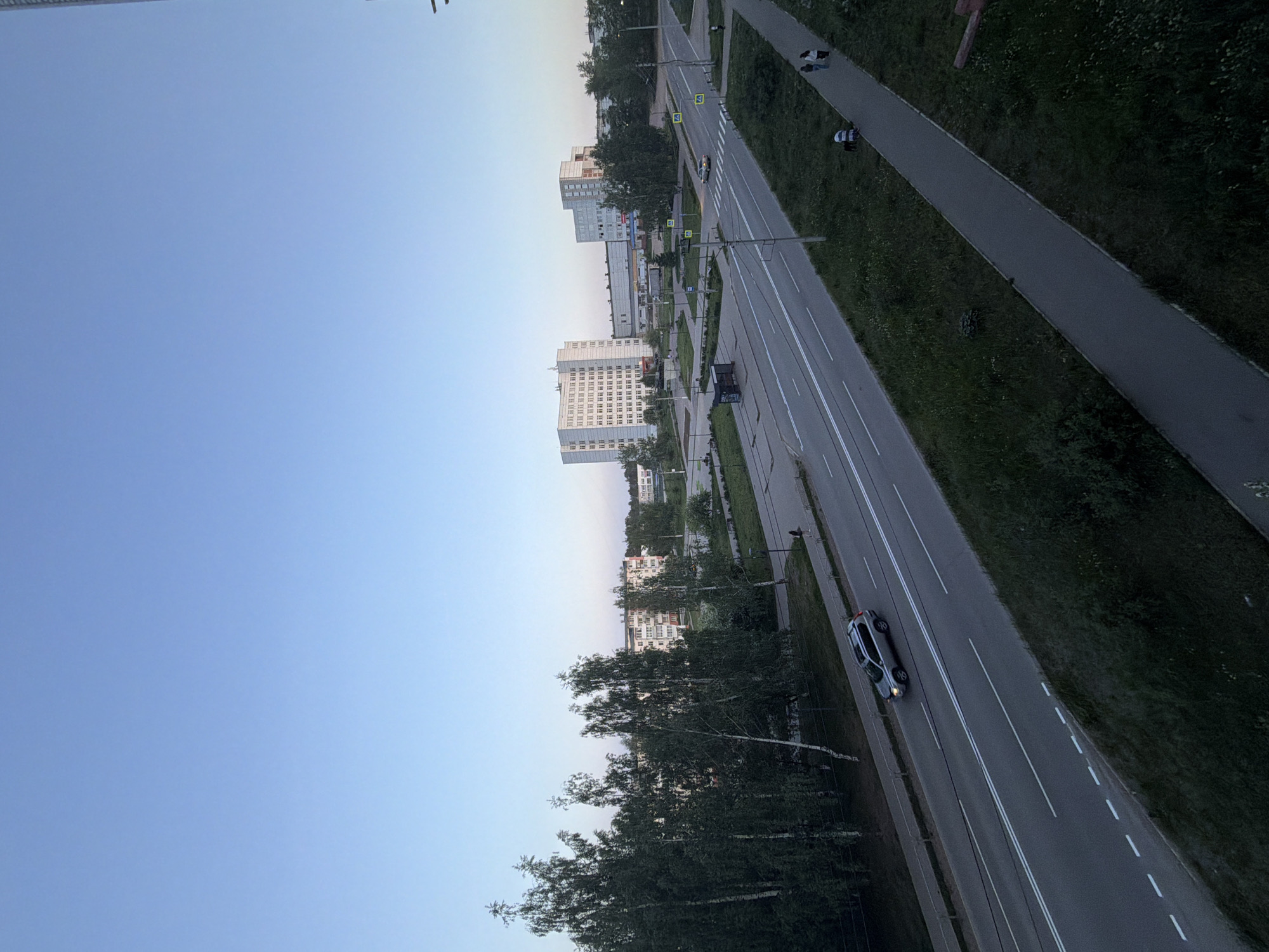 The image shows an aerial view of a street in the city of Chaikovsky, Russia. The street runs diagonally across the frame, with buildings visible in the distance. A car is visible on the street. The sky is blue. Trees line the side of the road. A pedestrian walkway is present next to the road.