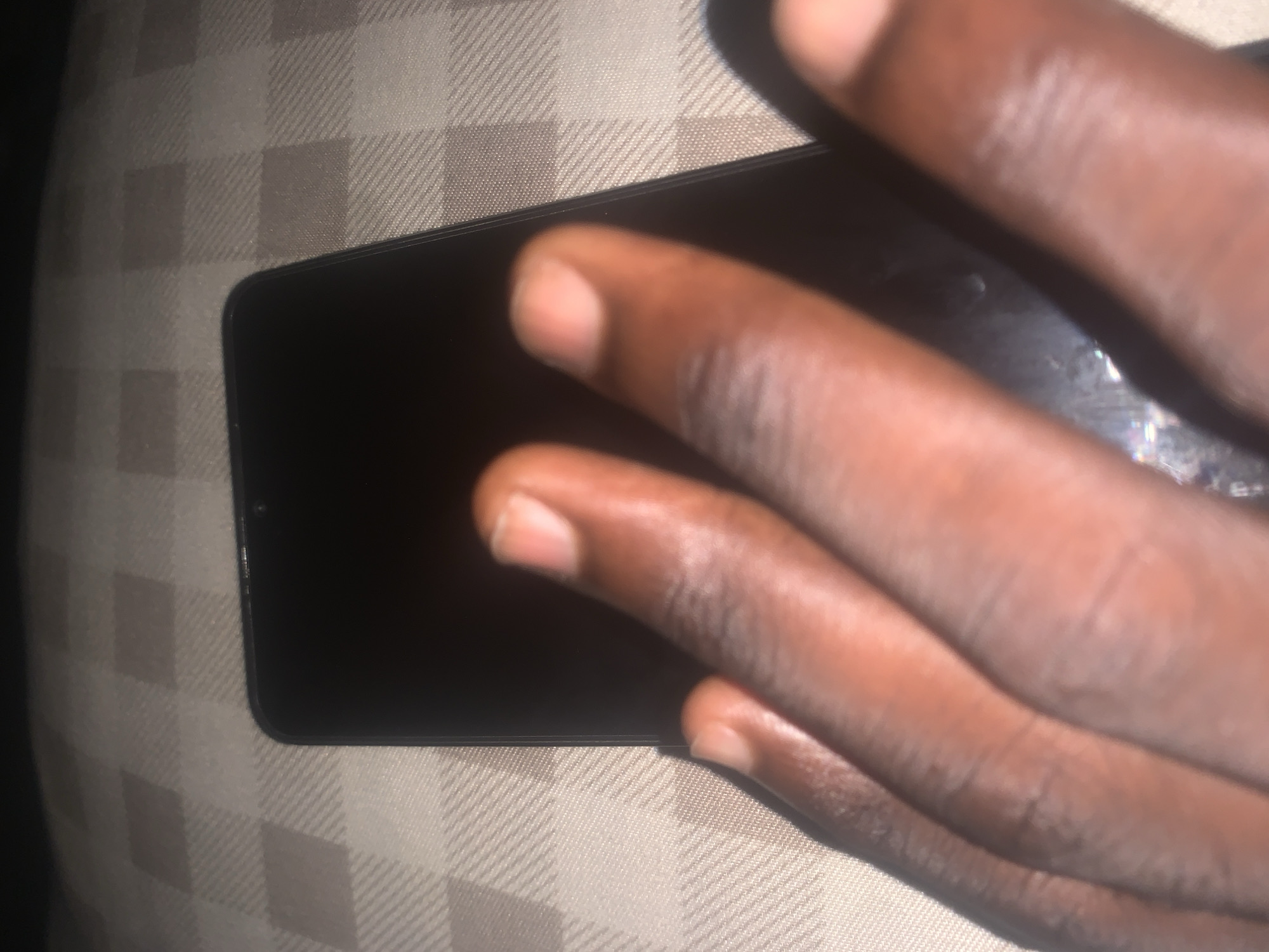 The image shows a close-up view of a person's hand resting on a dark-colored smartphone. The phone is placed on a beige and white checkered surface, possibly a tablecloth or bedspread. The hand, with visible fingerprints, covers a significant portion of the phone's screen. The lighting suggests an indoor setting, potentially at night or in a dimly lit room. The image does not provide any specific information about the location within Jalingo, Nigeria, as there are no identifiable landmarks or contextual clues. The focus is solely on the hand and the phone.