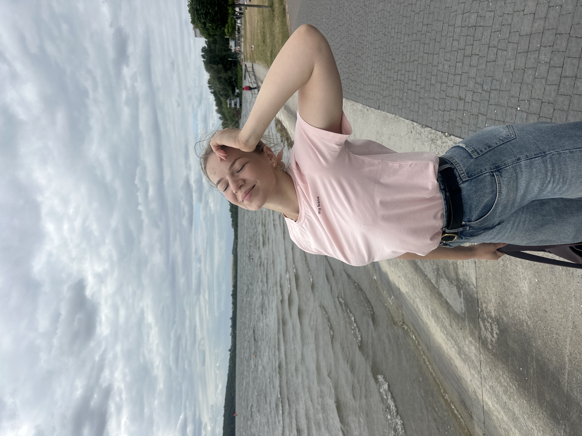 The image depicts a young woman standing on a paved promenade next to a body of water. The woman is wearing a light pink t-shirt with the text "my base" printed on it, and baggy blue jeans with a black belt. She has her right arm raised, resting her hand on her forehead, and her eyes are closed as if enjoying the moment. She is holding a dark-colored bag in her left hand.
The promenade appears to be a waterfront walkway, with grey paving stones. To the left of the promenade is a concrete embankment leading down to the water, which has small waves. Beyond the water, a distant treeline and buildings are visible. The sky is overcast with a mix of grey and white clouds.
The location is a waterfront promenade in Ternopil, Ukraine. Specifically, it is likely along the Ternopil Lake, a popular recreational area within the city. The presence of the paved walkway, embankment, and the expanse of water are characteristic of the lake's shore.