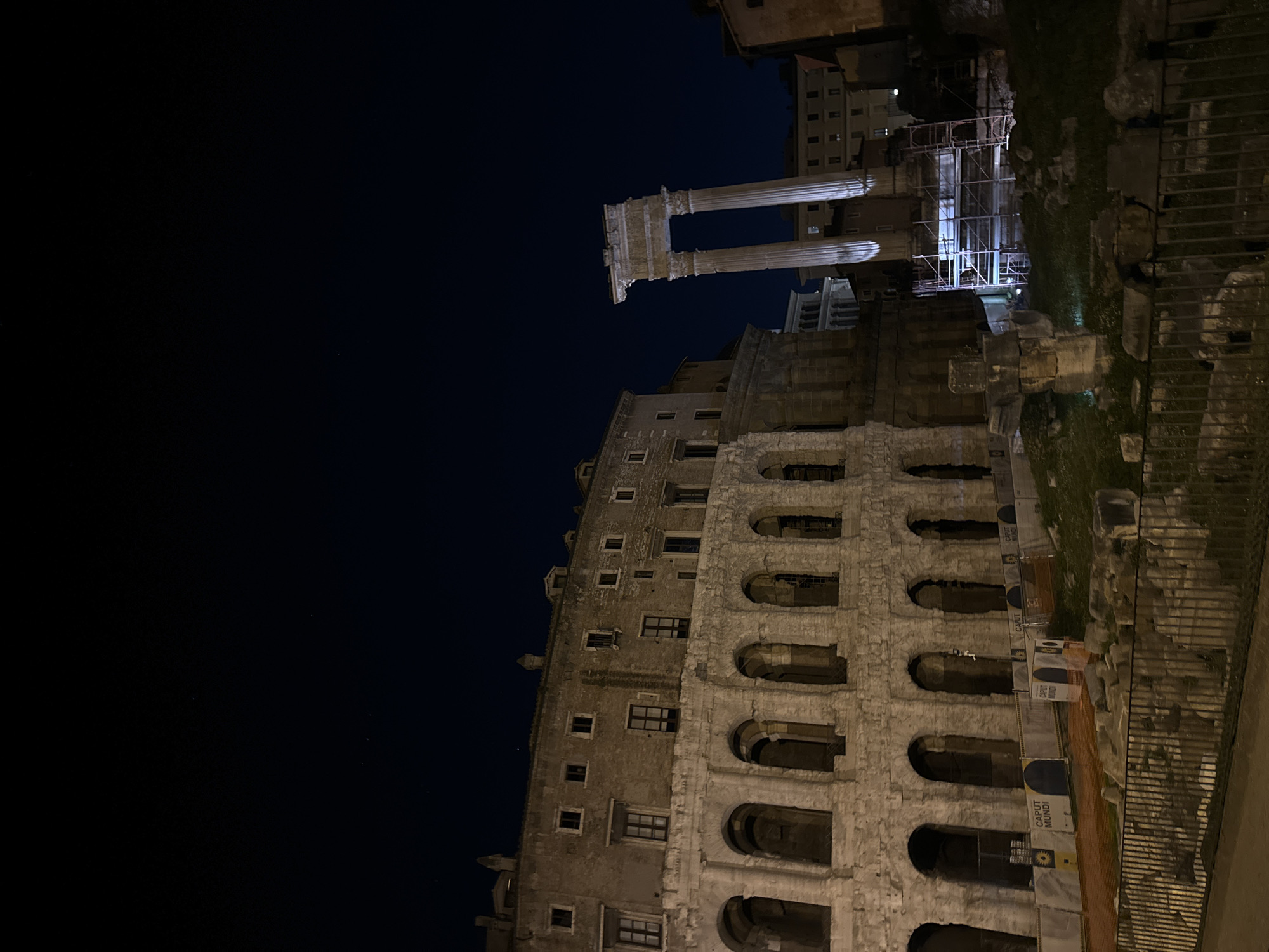 At night, the ruins of the Theatre of Marcellus in Rome, Italy, are illuminated. Adjacent to the theatre, the columns of the Temple of Apollo Sosianus are visible. Scaffolding surrounds part of the temple. The buildings are made of stone and feature multiple arches and windows. A dark sky is overhead. A fence appears in the foreground.