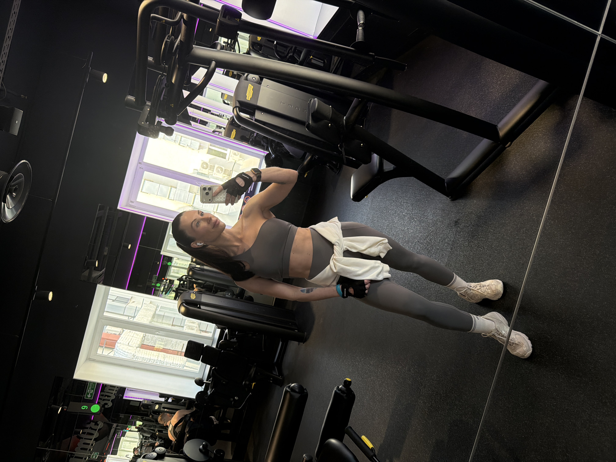 A woman in athletic wear (gray leggings and sports bra with a white sweatshirt tied around her waist) is taking a selfie in a gym. She is holding a smartphone in her right hand. The gym equipment in the background includes weight machines. Windows with daylight are visible. The image is likely taken in the city of Perm, Russia, given the location information.