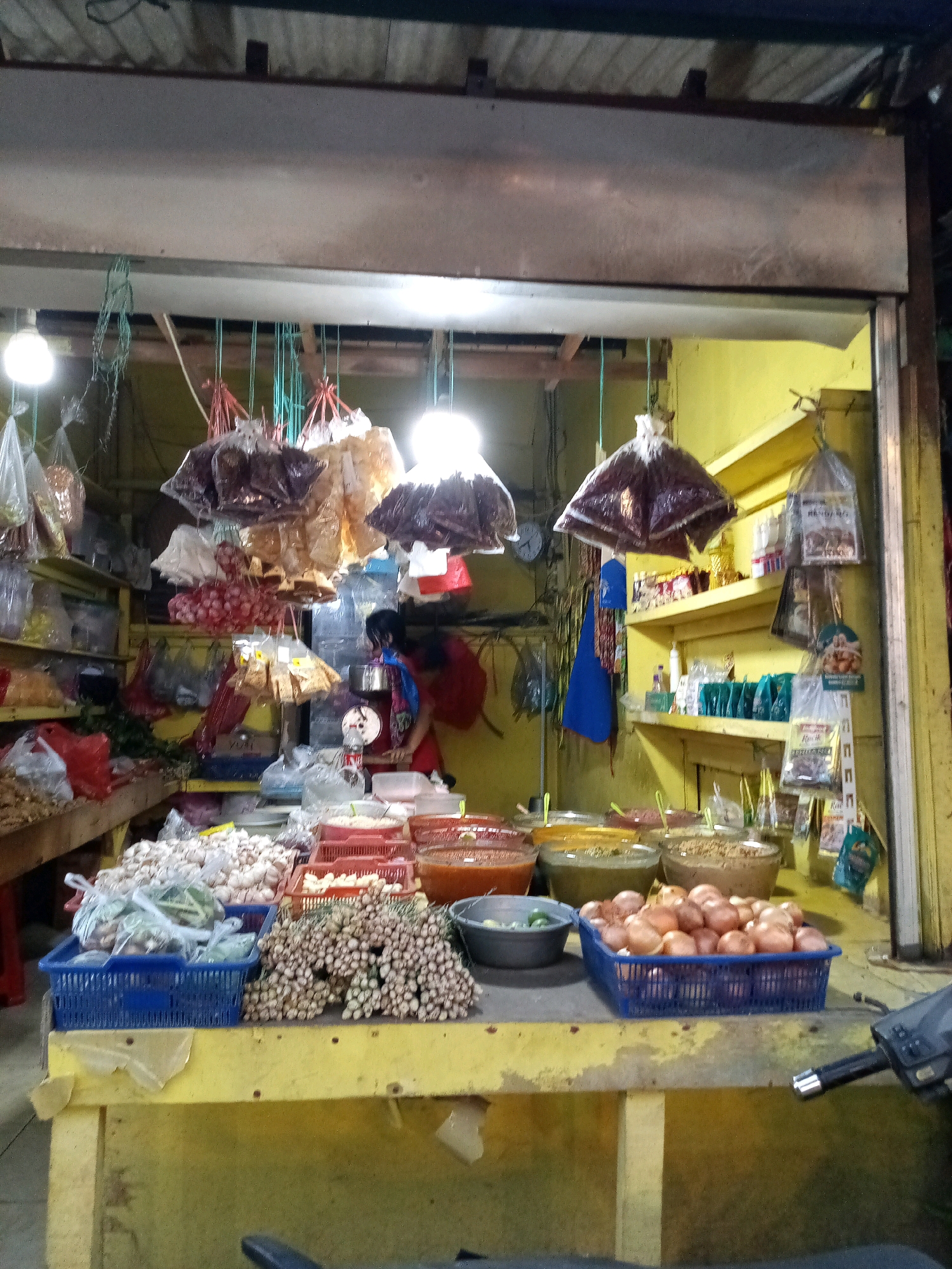 A modest outdoor market stall in Bekasi, Indonesia, displays a variety of fresh produce and packaged goods under bright fluorescent lights. The stall features a yellow wooden counter laden with goods. Piled high on the counter are bunches of small onions, white onions, and green garlic. To the right, a blue plastic basket overflows with red onions. Behind these, several large bowls contain what appear to be sauces or pastes in red, green, and brown hues. Shelves behind the counter are stocked with various packaged items, including bags of spices or dried goods, some in clear plastic and others in colorful packaging. Several bags of produce are also hung from the stall's overhead structure. A woman, partially visible, is attending to the stall. The overall scene depicts a typical local market offering everyday necessities to the community.
