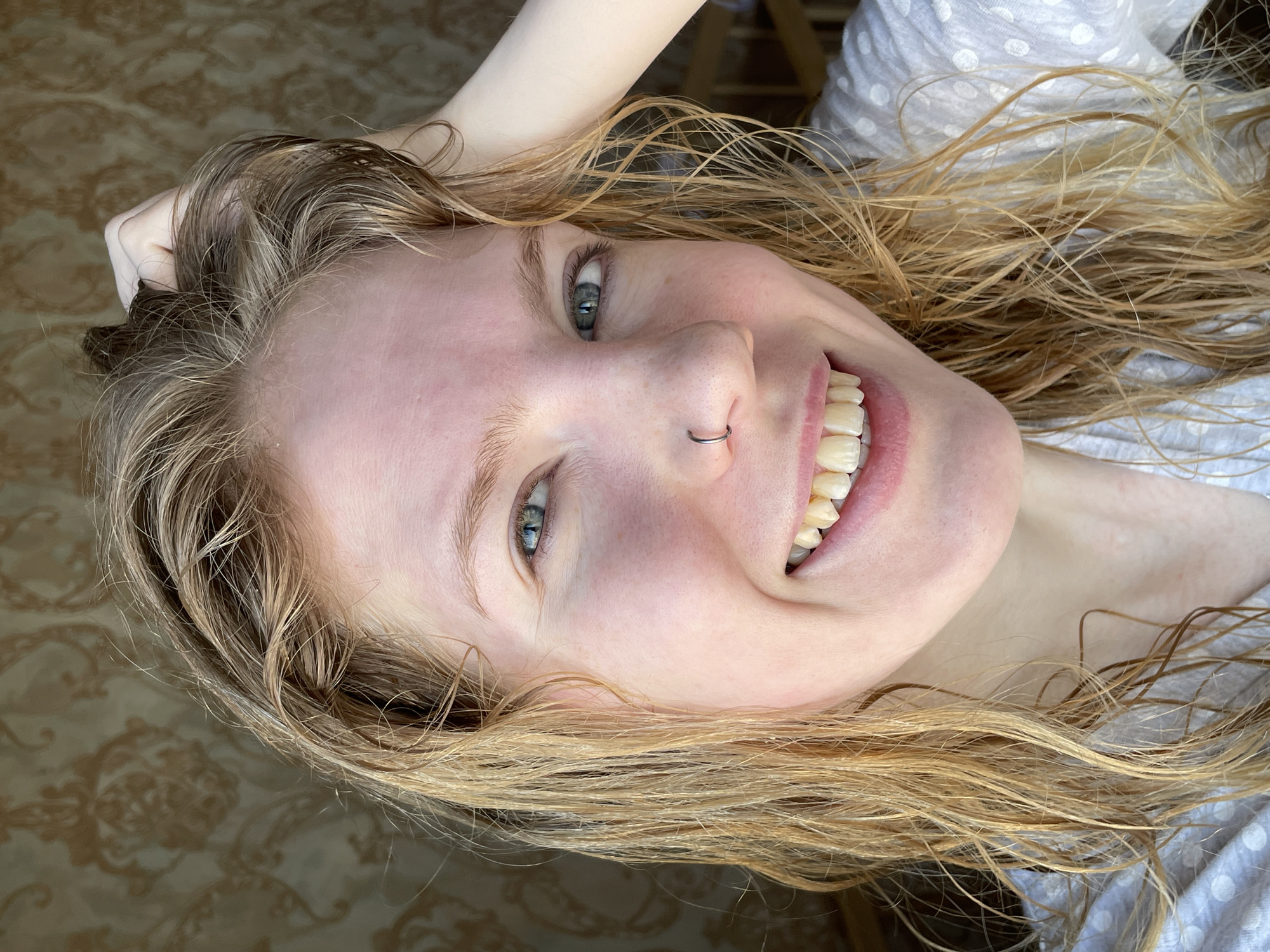A close-up, slightly rotated, high-angle shot shows a woman with blonde, shoulder-length wet hair, holding a portion of her hair in her left hand, with a visible silver nose ring on her left nostril. The woman is smiling, revealing her teeth. She has fair skin and blue eyes. She is wearing a white shirt with light gray polka dots. The background consists of a patterned surface and a blurred wooden chair. There is no indication of the location of Izhevsk, Russia.