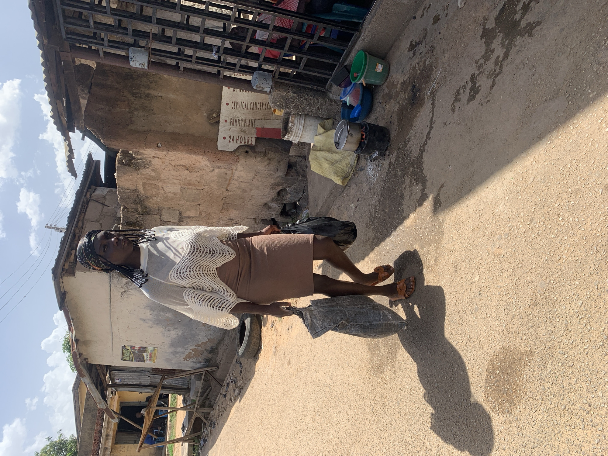 A young woman stands on a paved street in Jos, Nigeria. She is wearing a tan, bodycon dress, sandals, and a white crochet top. She also has braids. She holds a large grey bag in her right hand. Behind her is a weathered, multi-story building with a sign indicating "Cervical Cancer Screening" and "Family Planning", with "24 hours" written below. A collection of buckets and containers are stacked near the building's base. The street is wet in places, reflecting the sunlight.