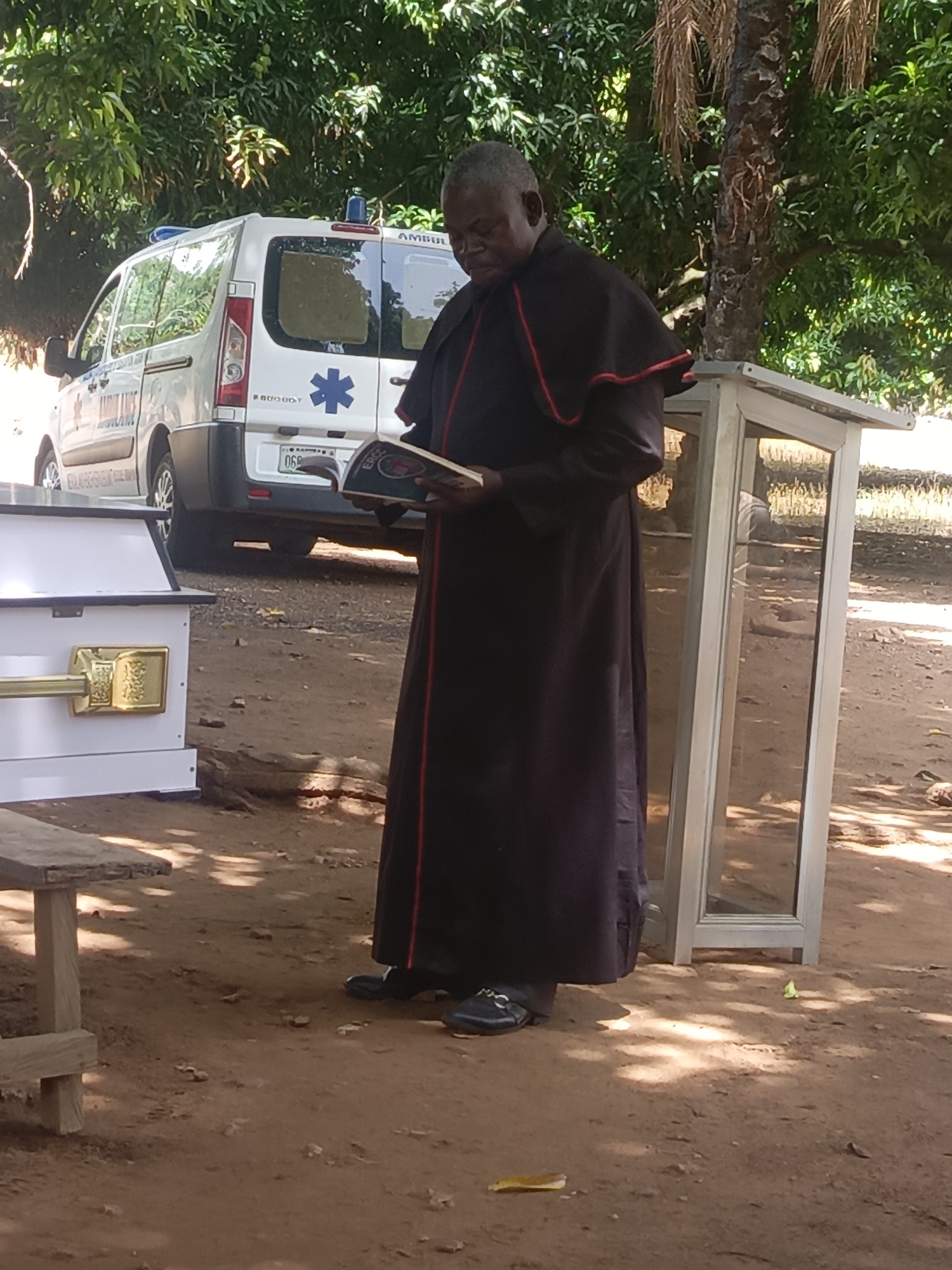 At an outdoor location near Ninzo South, Nigeria, a funeral ceremony is taking place. An adult male in religious attire reads from a book. A white coffin is visible to the left of the male, while a white ambulance is visible in the background. The area is dirt or soil, with scattered leaves on the ground. The setting includes trees and an open structure with a reflective surface, possibly glass.