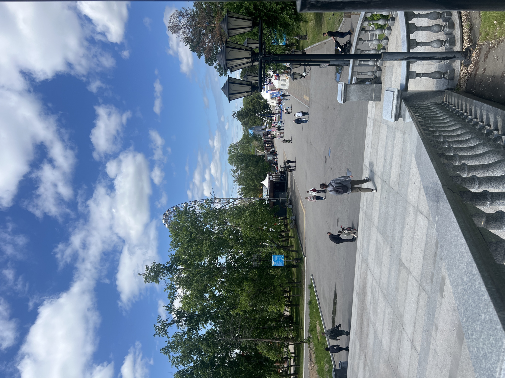 This image depicts a vibrant daytime scene in Dynamo Park of Culture and Rest (Парк культуры и отдыха «Динамо») in Khabarovsk, Russia. The perspective is from an elevated position, looking down a wide, paved main thoroughfare that extends into the distance.
In the immediate foreground, a gray granite balustrade with multiple decorative balusters is visible, extending from the bottom right towards the center-left. A large section of light gray, rectangular tiled pavement occupies the foreground, leading up to the main pathway.
The midground is dominated by a wide, dark gray paved path or road, possibly asphalt, which has some lighter patches that may be shallow puddles or reflective areas. Numerous people are present on this path. On the far left, two individuals (one in a dark jacket, one in a lighter jacket with a backpack) walk away from the viewer. Towards the center-left, a man in dark attire is bending over, assisting a small child on a balance bike or similar small wheeled toy. The child is wearing light-colored clothing. To the right of the child, a person with a light-colored backpack walks away. On the far right, another person in a dark shirt pushes a baby stroller. Further down the path, various individuals and small groups are seen walking in both directions.
Lush green deciduous trees, featuring dense foliage, line both sides of the main pathway, indicating a spring or summer season. A taller, multi-lamp street light with decorative lantern-style fixtures stands on the right, partially obscured by the frame. On the left side of the path, a blue informational sign on a pole is visible near a dark trash receptacle. Further into the midground on the right, several white event tents or market stalls are set up along the path, and a tall, blue and white amusement ride structure, possibly a bungee trampoline, is visible.
In the background, a large Ferris wheel with a dark, open metal frame is prominent, partially obscured by trees on the left. The sky above is a bright, clear blue with scattered, fluffy white cumulus clouds. The overall scene suggests a busy park environment on a sunny day.