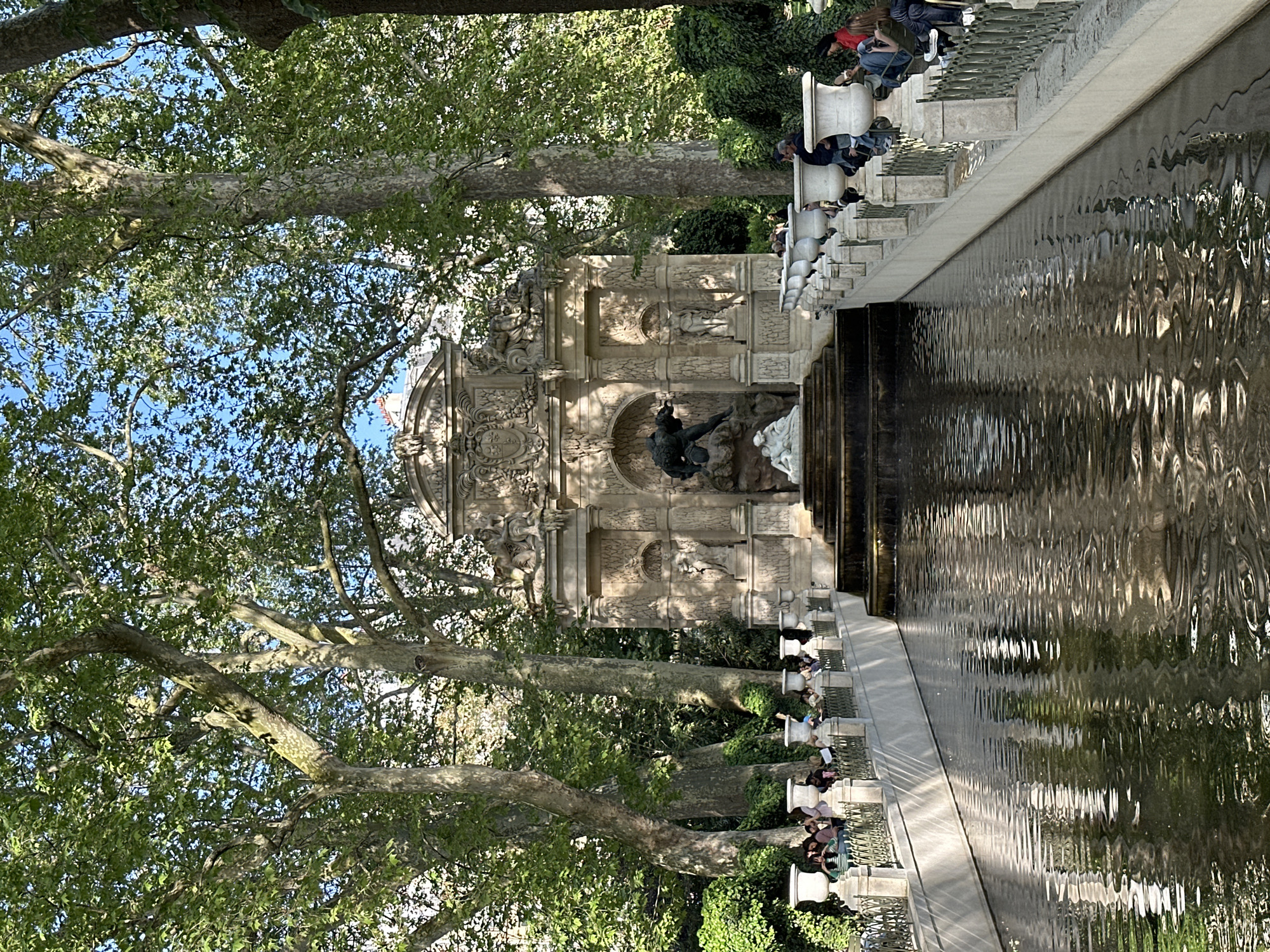 This image captures a serene scene at the Medici Fountain in the Jardin du Luxembourg, Paris, under a bright, clear sky. The prominent subject is the ornate stone fountain structure, featuring classical sculptures, including figures within a grotto and a cyclops above, surrounded by intricate carvings. Extending from the fountain is a long, rectangular reflecting pool, its surface rippled and shimmering with reflections of light and the surrounding greenery.
The fountain and pool are framed by large, mature trees with distinctive mottled bark and lush green foliage, through which patches of blue sky are visible. Along the right side of the reflecting pool, a raised stone platform with a decorative balustrade offers numerous individual stone seats. Several people are scattered across these seats, engaging in leisurely activities such as resting, conversing, or observing the tranquil surroundings. Their casual attire suggests a relaxed daytime outing.
The time of day appears to be late morning or afternoon, indicated by the strong sunlight and well-defined shadows. The weather is pleasant and sunny, enhancing the peaceful and idyllic atmosphere of the garden. The architectural style of the fountain serves as a strong location cue, identifying it as a famous Parisian landmark. There is no discernible text visible in the image.