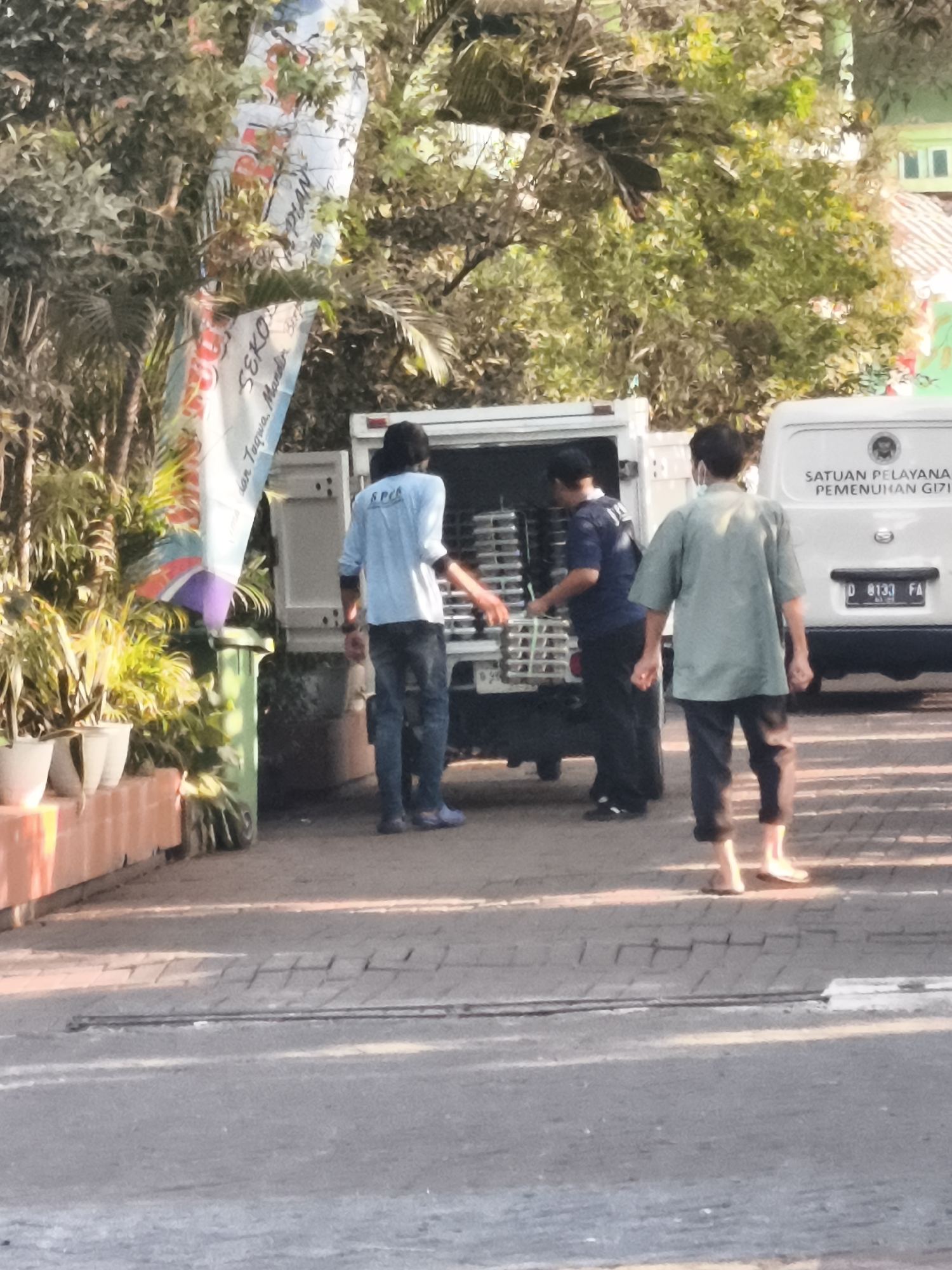 In North Jakarta, Indonesia, three men are near a white delivery truck with its back doors open. One man, wearing a light blue shirt and dark pants, is standing in the truck, arranging what appears to be stacks of food containers. Another man, in a dark blue shirt and dark pants, is partially visible inside the truck, also attending to the containers. The third man, in a light green short-sleeved shirt and dark pants, stands outside the truck, facing away from the camera, with his hands clasped in front of him. A white van with Indonesian license plate D 8133 FA and text reading "SATUAN PELAYANA PEMENUHAN GIZI" is parked behind the truck. In the foreground, a patterned walkway leads to a street. Lush green foliage and trees surround the scene, with a tall banner featuring Indonesian text and illustrations hanging from a tree to the left of the truck. Several potted plants are arranged in a row along a red brick border in the lower left corner.