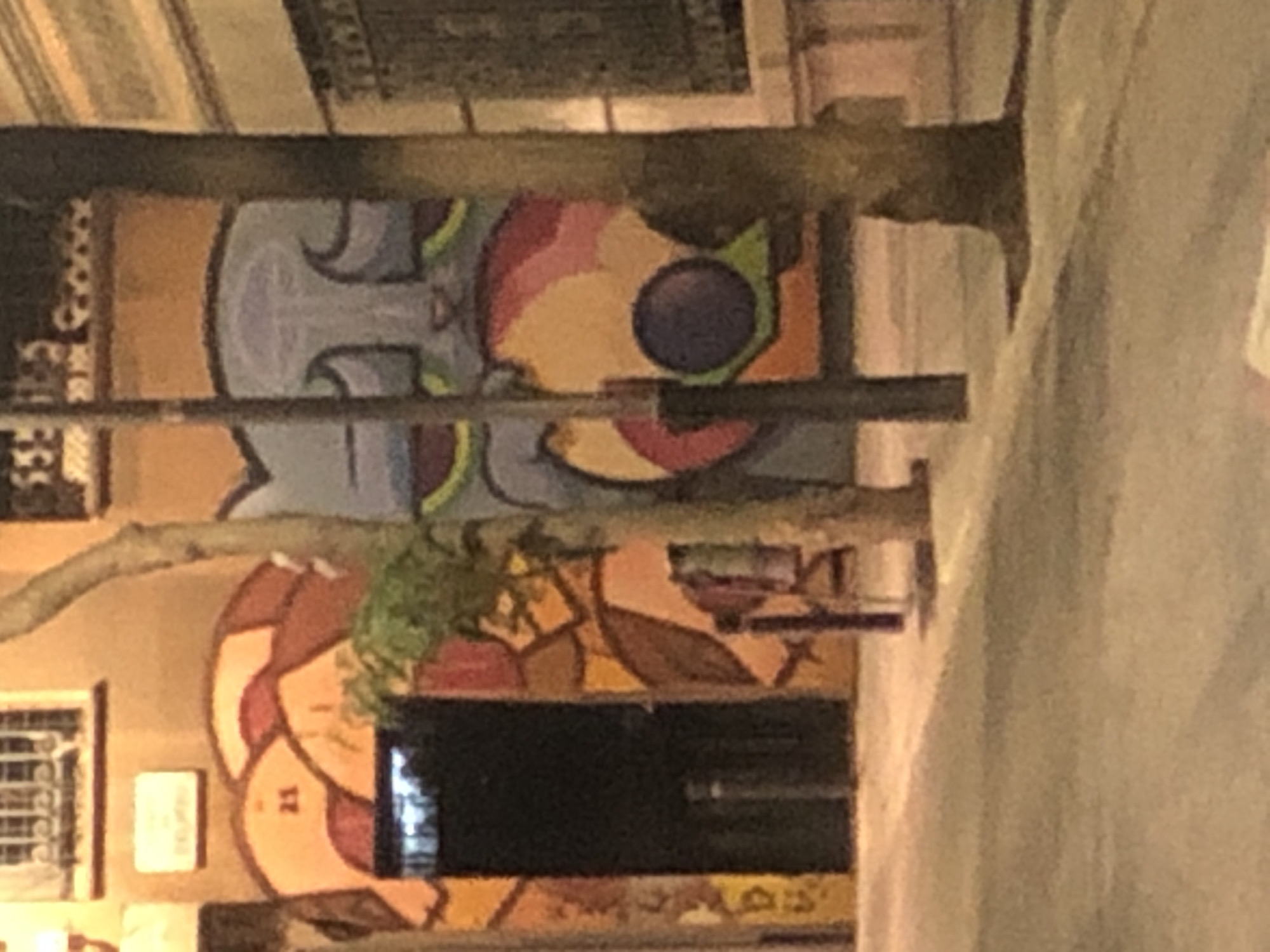 The image shows a street scene with graffiti art on a building. The graffiti depicts a large, stylized cat face with blue and purple tones. To the left of the cat, there is an arched doorway with colorful geometric patterns and the number "21" is visible on the wall above the arch. Trees with rough bark are growing in front of the building, partially obscuring the graffiti. A lamppost stands in front of the cat mural. The street in the foreground is paved and appears to be dimly lit, suggesting it might be nighttime. The buildings in the background have windows and architectural details.