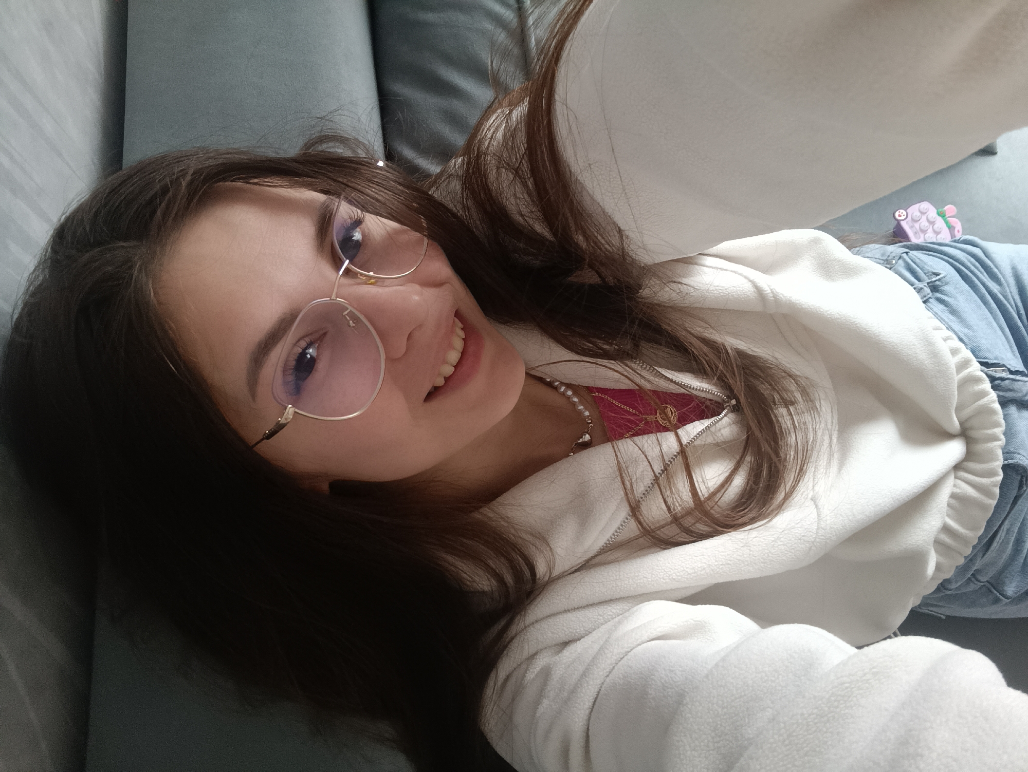 A young woman with long brown hair, wearing gold-rimmed glasses, a white hooded sweatshirt, a pink and red top, and blue jeans is lying on a gray upholstered surface, possibly a sofa. A small purple object is visible on her jeans. The location is Unknown, Unknown.