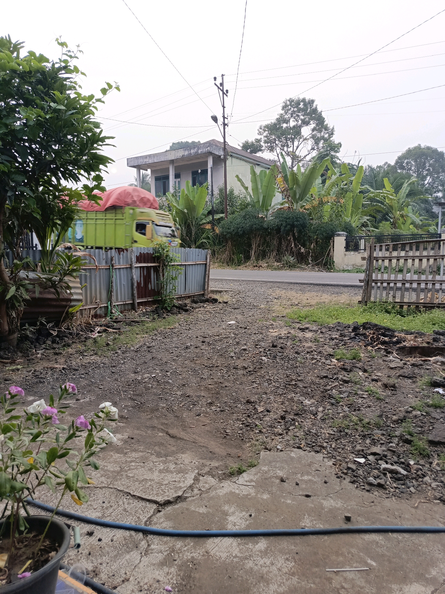 In Ulak Tanding, Indonesia, a cloudy day is depicted with a truck covered in a red tarp driving past a corrugated metal fence. Behind the fence, a house with a modern design stands amidst lush greenery and banana trees. The foreground features a cracked concrete surface with a black pot containing flowering plants and a blue hose. The ground is covered with gravel and debris, with a wooden fence visible to the right. The scene captures a rural or suburban setting.