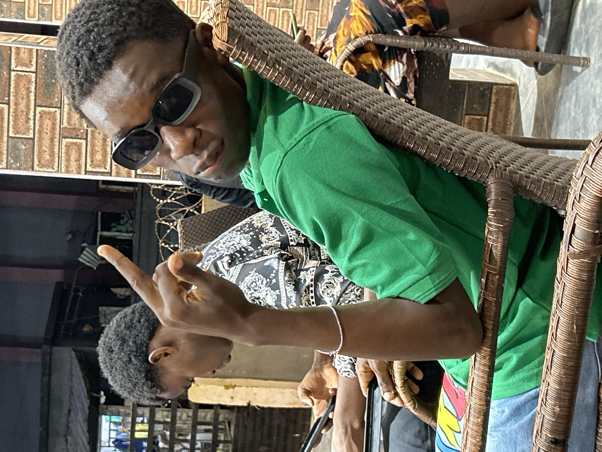 The image captures a close-up of two young men sitting on woven chairs outdoors, likely in Ihiagwa, Nigeria, during daylight hours. The man in the foreground, wearing a bright green t-shirt and sunglasses, is leaning back and holding his left hand up, fingers extended in a gesture. His right arm rests on the chair. To his left, another young man, wearing a patterned shirt, is seated and looking down at a mobile phone in his hands. The background shows a brick wall and some architectural elements of what appears to be a commercial or residential building, with a wire fence visible. The ground is tiled. The overall setting suggests a casual, outdoor gathering.