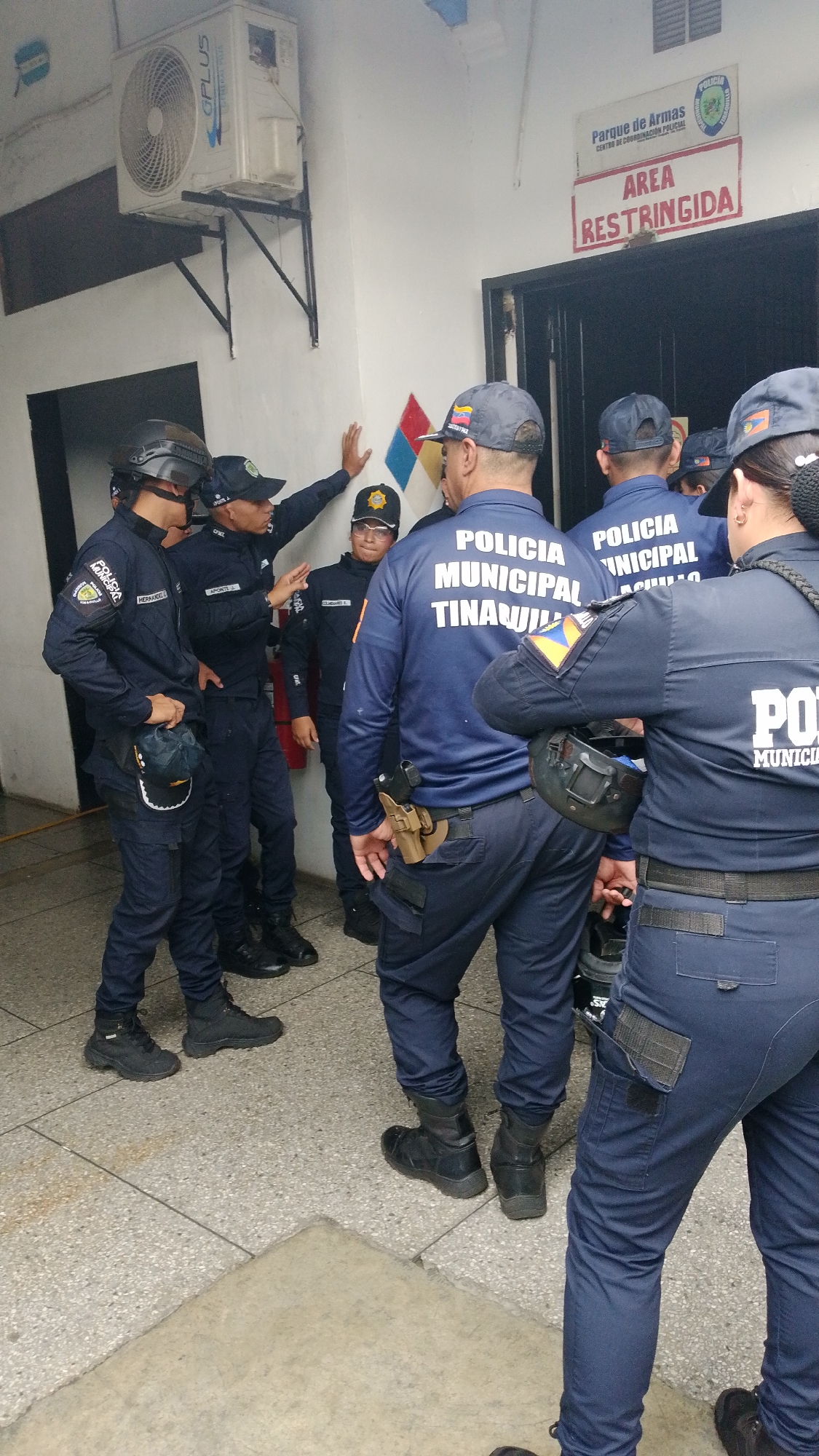 A group of municipal police officers from Tinaquillo, Venezuela, stand in a semi-enclosed area marked as restricted. The officers wear dark blue uniforms with the words "POLICIA MUNICIPAL TINAQUILLO" on their backs. Some have firearms holstered at their waists. The environment includes a tiled floor, a wall-mounted air conditioning unit, and a sign indicating a restricted area, possibly an armory ("Parque de Armas"). One officer gestures, seemingly providing instructions or information.