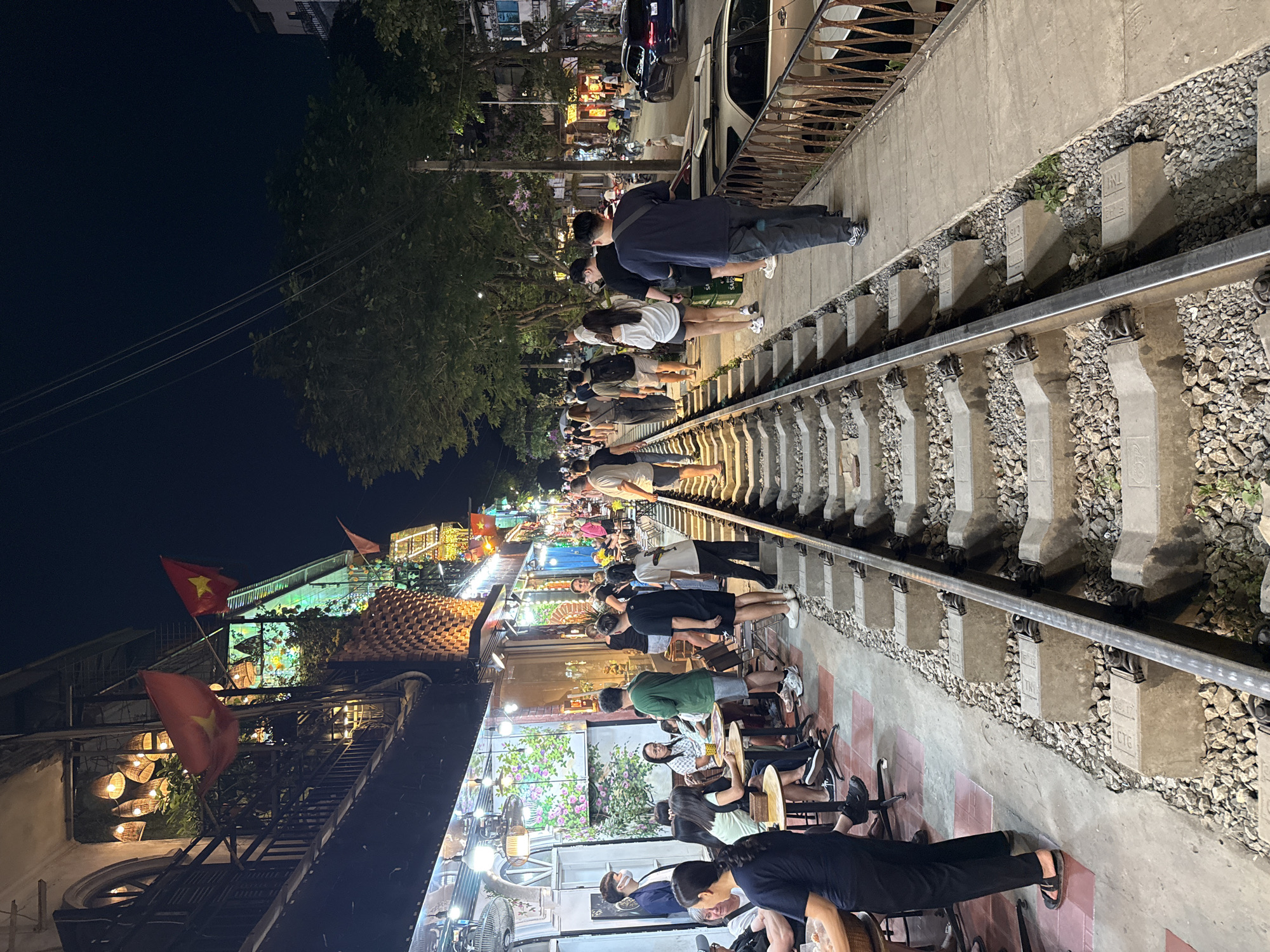 The image captures a lively nighttime scene along railway tracks in an urban setting, likely Hà Nội, Vietnam. The tracks run down the center of the frame, flanked on both sides by concrete ground and, further back, by buildings that house cafes and restaurants.
Numerous people, mostly young adults, are gathered along both sides of the railway. Many are standing close to the tracks, some appearing to observe the surroundings or socialize. Further back on the left, more people are seated at tables and chairs within brightly lit establishments, suggesting dining or leisure activities. The atmosphere appears social and bustling, illuminated by string lights, lanterns, and various storefront lights from the businesses lining the route.
Notable details include several red Vietnamese flags with yellow stars, which are prominently displayed, especially on the left side of the image, affirming the location context. Green trees and foliage are visible above and behind the buildings, with a dark night sky overhead, occasionally intersected by power lines. Cars and motorbikes can be seen parked or moving on a street parallel to the tracks in the background.
The railway itself consists of metal rails resting on concrete sleepers, which are embedded in gravel. Some of these concrete sleepers have text markings, including "TN1," "HSN," "CT0," "CTQ," and numbers such as "08" or "98," likely identifying railway infrastructure. The concrete walls and barriers alongside the tracks show signs of age and greenery growing in crevices. A black fan is visible in the foreground area of one of the cafes.