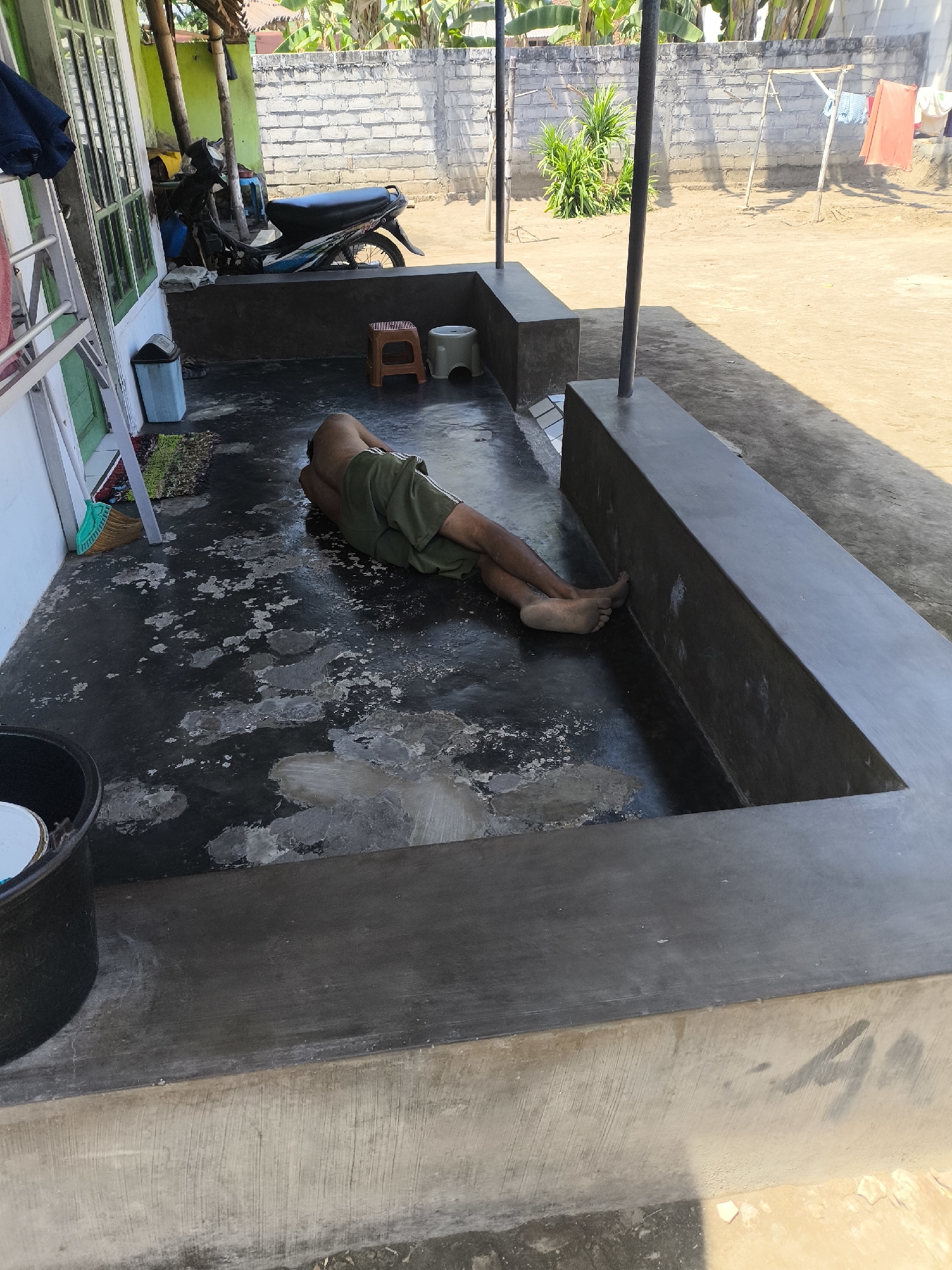 A person, dressed in green shorts, is sleeping on their side on a dark, worn concrete patio. The patio is partially enclosed by a low, dark concrete wall. The person is positioned with their head and torso within the enclosed area, and their legs extend out onto the patio floor. Their feet are bare and dirty. To the left of the person, there is a white wall with green-trimmed windows and a metal ladder leaning against it. A blue trash can and a broom with a green head are also visible near the wall. A motorcycle is parked to the left of the patio, near a building with a thatched roof. In the background, a light-colored brick wall surrounds an outdoor area with a clothesline and some plants. The ground in the background appears to be dirt or sand. The scene is illuminated by bright sunlight, casting shadows. The overall impression is that of a simple, outdoor living space in a rural or semi-rural Indonesian setting, possibly Grati, Indonesia, given the architectural style and vegetation.