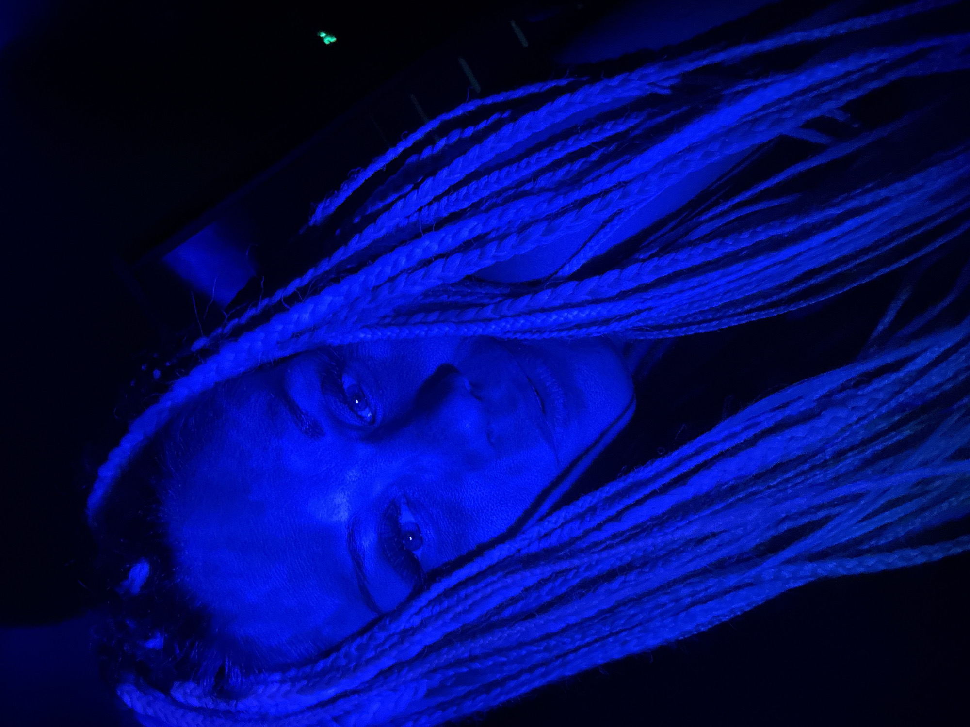 A close-up, over-the-shoulder shot shows a person's face illuminated by blue light. The person has blonde hair styled in numerous braids that fall over their shoulders and face. Their eyes are open and looking towards the camera, with visible eyeliner. The background is dark, with some faint green lights visible in the upper right quadrant of the image, suggesting an indoor environment. There is no information in the image that indicates a specific location in the city of Rusanovo, Russia.