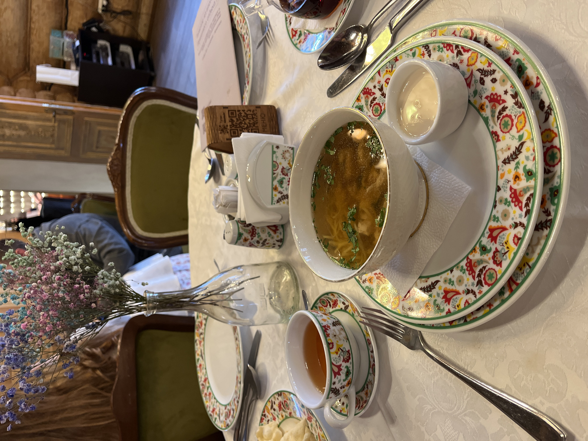 The image depicts a table setting at a restaurant, likely in Kazan, Russia, given the traditional floral patterns on the tableware. A bowl of soup is prominently featured on a patterned plate, accompanied by a small bowl of cream. Nearby, there is a teacup with tea and a plate with small dumplings. Silverware is laid out beside the plates. A small vase with a colorful bouquet of flowers sits on the table, near a wooden holder with napkins and condiments. In the background, one can see a portion of a decorative, possibly wooden, wall and a blurred figure seated in a green chair. The overall setting suggests a cozy, traditional restaurant atmosphere.