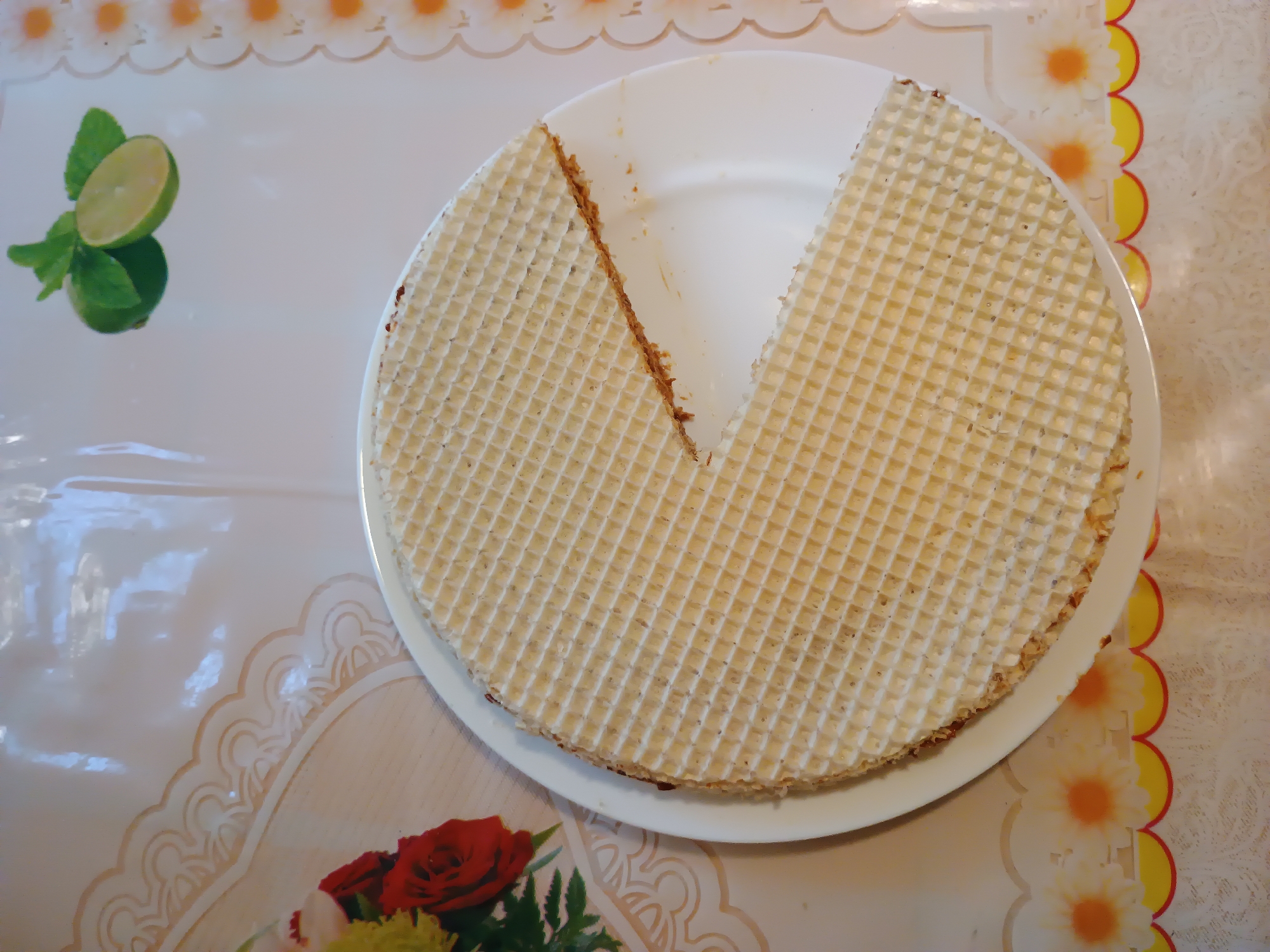 A circular, layered wafer cake sits on a white plate, partially sliced, revealing a thin layer of filling. The cake is positioned on a white tablecloth with a decorative scalloped border featuring floral motifs. To the left of the plate, a slice of lime with two green leaves is visible, also against the patterned tablecloth. In the lower left corner of the image, a bouquet of red roses and greenery is partially visible. The scene is brightly lit, suggesting daylight. The overall setting appears to be a domestic environment, possibly a kitchen or dining area, in Zaporizhzhia, Ukraine. No text is visible in the image.