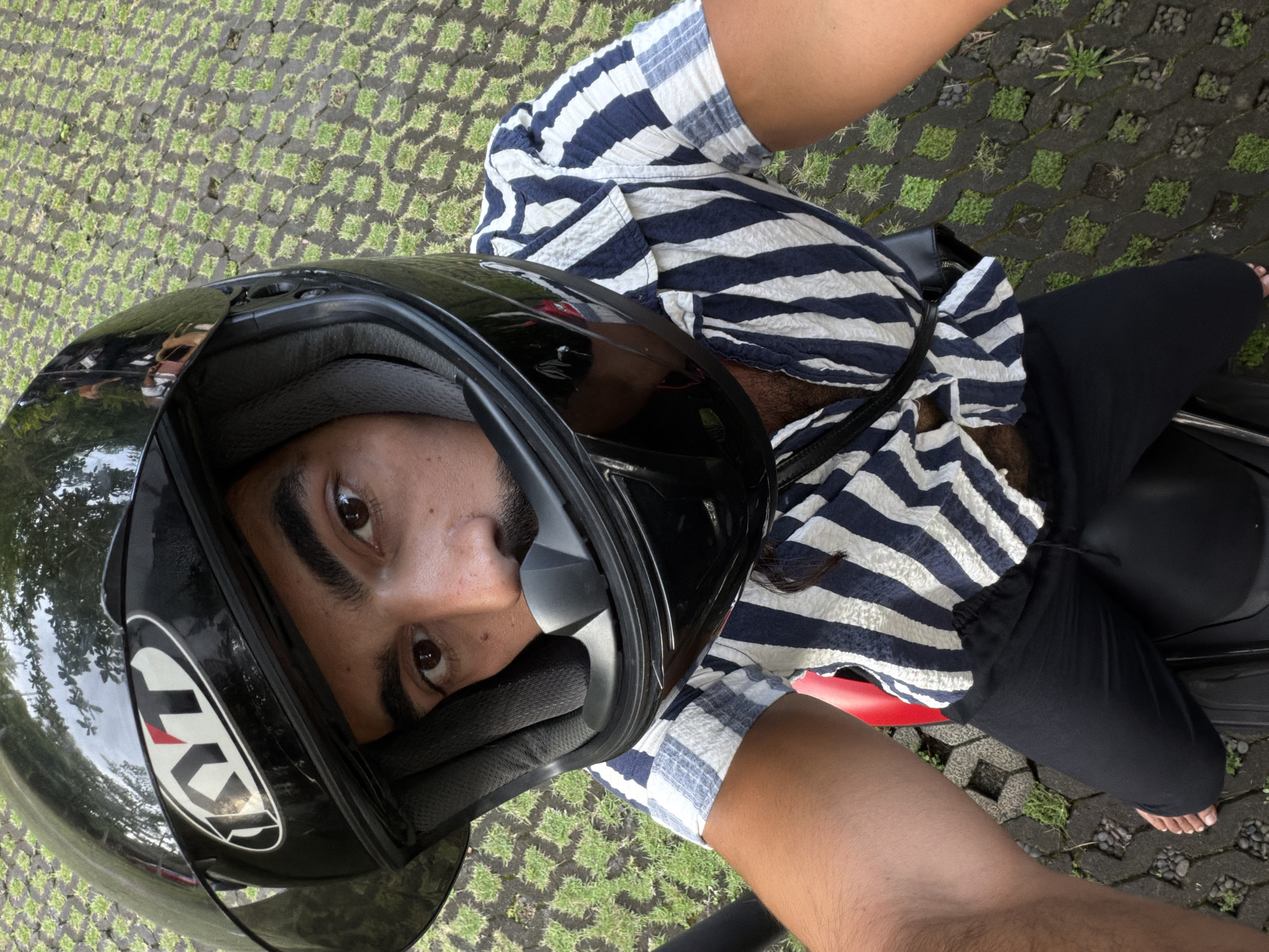 A person wearing a black motorcycle helmet and a striped, button-down shirt takes a selfie from a high-angle perspective. The person is seated on a red motorcycle with black pants. The ground surface is made of interlocking pavement blocks with grass growing between them. The location is determined to be Sukawati, Indonesia based on the context.