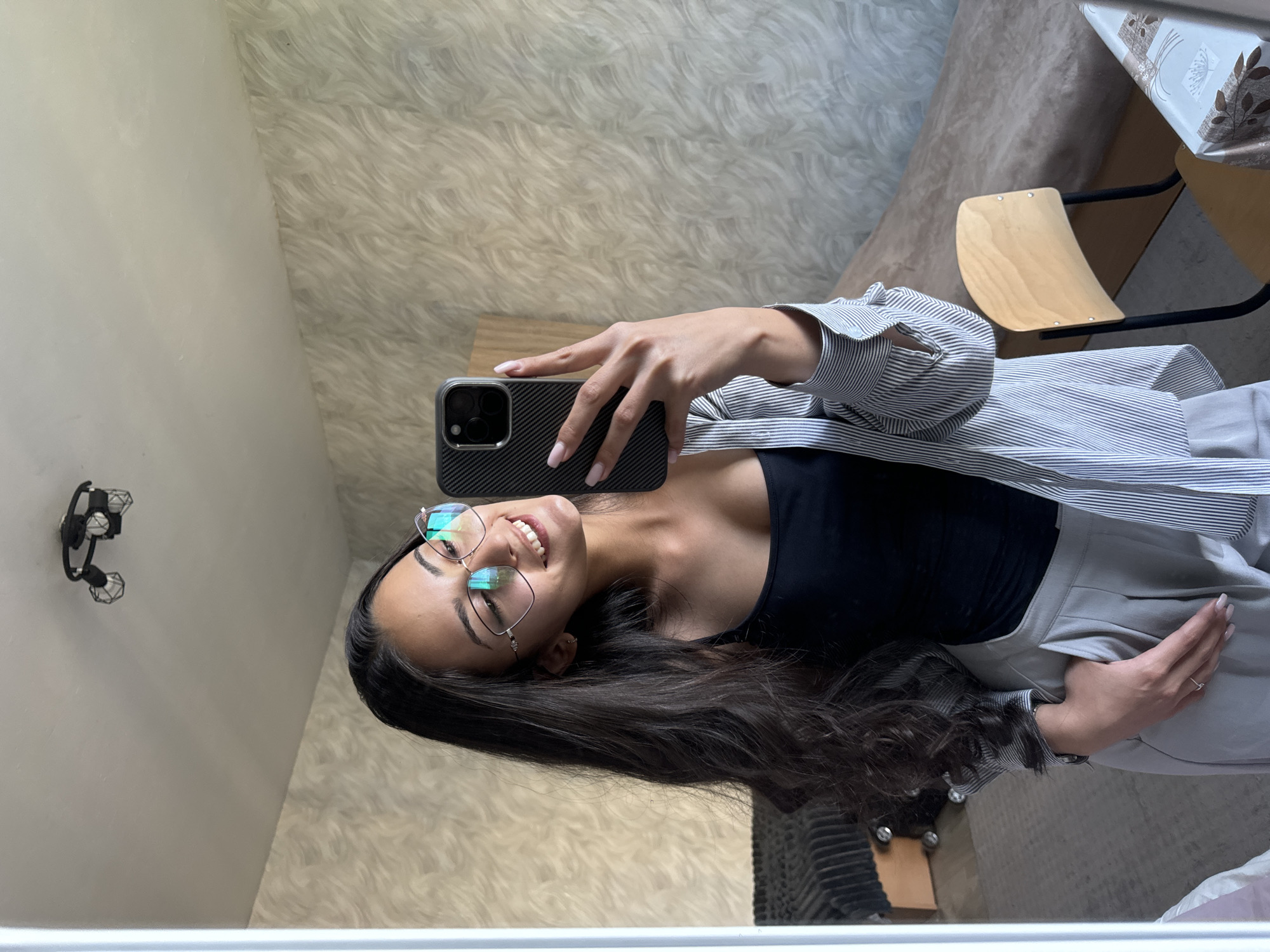 A woman is taking a selfie in front of a mirror. She is wearing glasses, a black top, a striped shirt, and light-colored pants. She is holding a smartphone with a dark case. A wall sconce is visible on the left side of the mirror. The background shows a patterned wall, a wooden table, and a chair with a wooden seat and black legs. The location is not discernible from the image.