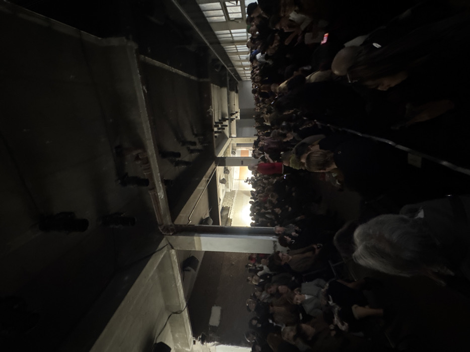 The image displays a densely attended indoor event within an industrial-style building. Hundreds of individuals are seated in rows on simple chairs, facing a brightly illuminated area at the far end of the elongated room, suggesting a stage or presentation space. The venue features exposed concrete pillars and walls, a dark ceiling with visible pipes, ducts, and numerous black, cylindrical track lighting fixtures. Natural light filters in through a series of multi-pane windows along the right-hand wall. Artificial lighting within the seating area is subdued, while the distant focal point is brightly lit. A camera on a tripod is visible in the background's illuminated section, indicating recording activity. Most attendees are dressed in dark attire, with one individual prominent in a red garment near the center-back. The specific location within Paris, France, cannot be determined from the visual evidence presented.