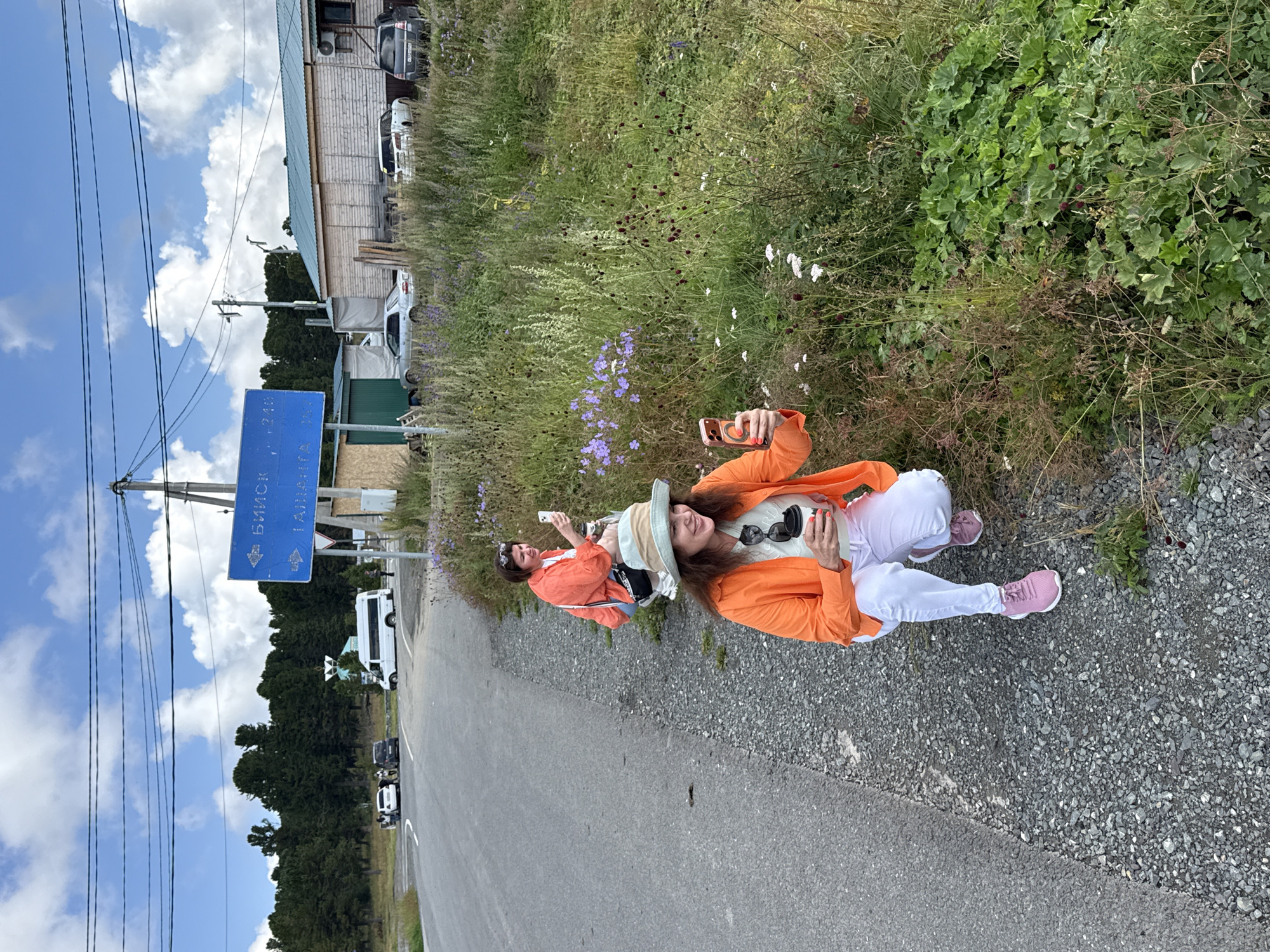 In T/B Seminskii Pereval, Russia, two women are captured in a roadside scene. The woman in the foreground, wearing an orange jacket, a light-colored hat, white pants, and pink sneakers, is squatting near a patch of overgrown grass and wildflowers. She is holding a disposable coffee cup and her phone, which is angled towards the flowers. Behind her, a second woman, also in an orange jacket and jeans, squats closer to the road, holding her phone up as if taking a picture. The road, composed of asphalt and gravel, curves to the left. A blue directional sign with Russian text indicates directions and distances. A cluster of trees forms a backdrop to the scene, with a few buildings and parked vehicles visible in the distance under a cloudy sky.