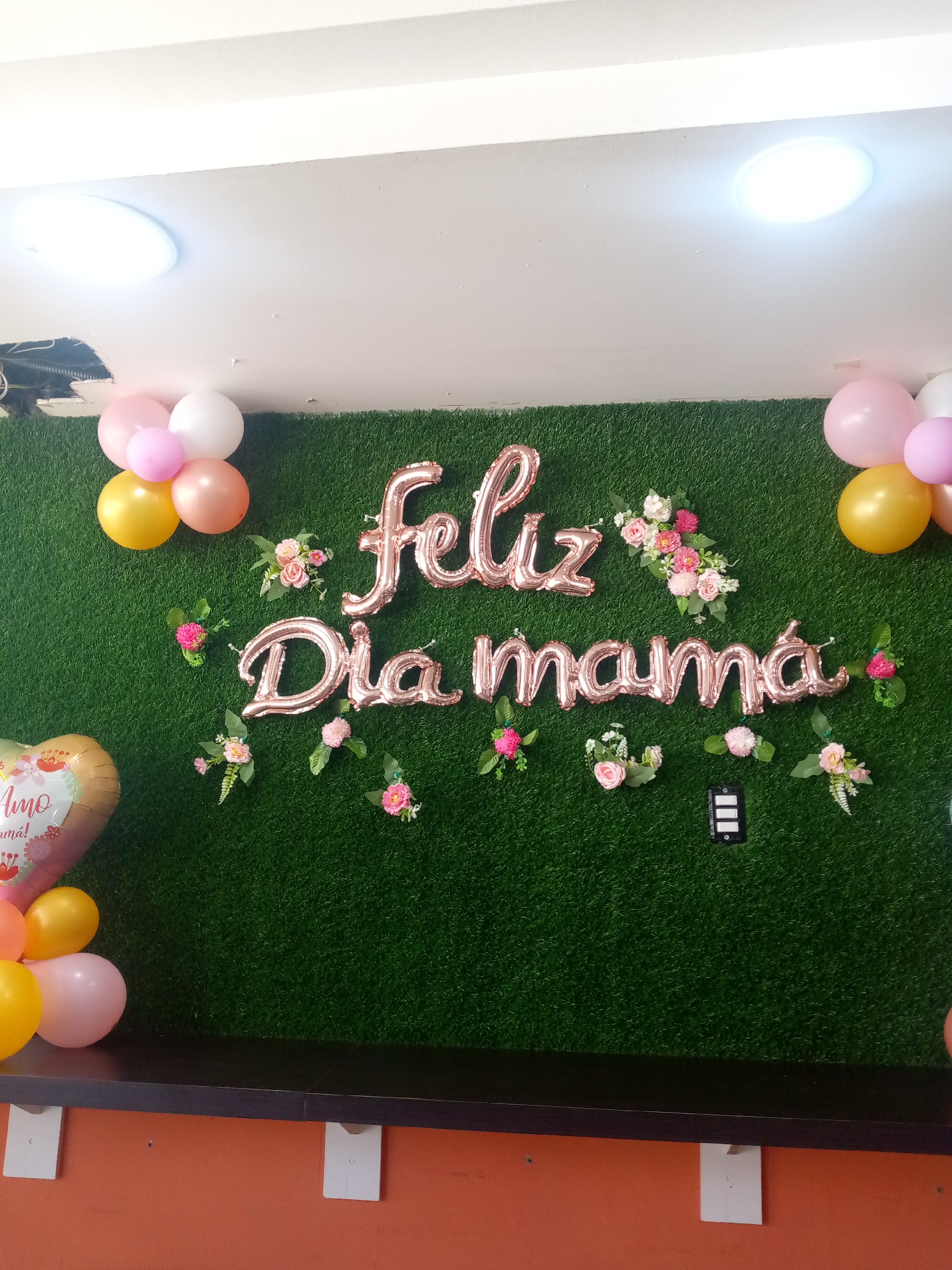 The image shows an indoor scene, likely a decorated event space. The background is a wall covered in artificial green grass. On the wall, there are rose gold foil balloons spelling out "Feliz Día Mamá" (Happy Mother's Day). The decorations include small bouquets of pink and white flowers arranged around the balloon words. Balloons in shades of pink, white, and gold are clustered on either side of the decoration. A black shelf runs along the bottom of the wall. The lower part of the frame shows the wall below the shelf is painted in a terracotta color. There is a balloon with the words "Te amo mam!" (I love you mom!) on the lower left of the display. The location of the scene is Unknown, Unknown.