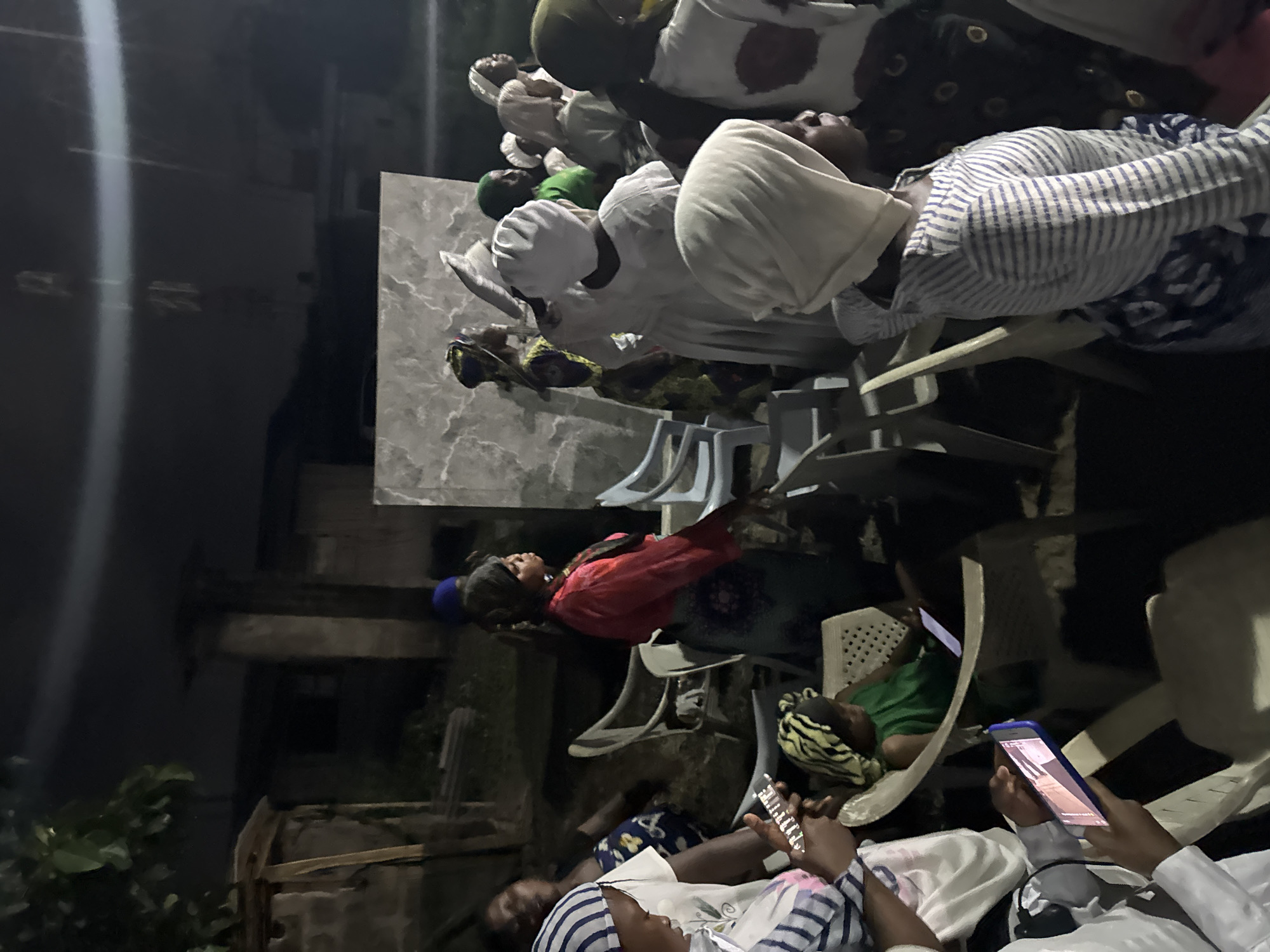The image captures a night scene in Badagry, Nigeria. A group of people are seated on plastic chairs outdoors. Some individuals are wearing white head coverings and light-colored clothing, while others wear colorful dresses and headscarves. A few people are looking at their mobile phones. The setting appears to be a residential area, with a building visible in the background.
