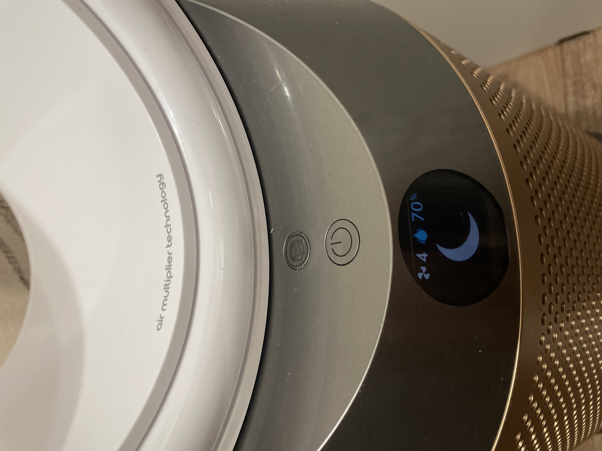 A close-up, eye-level, indoor shot shows the top portion of a Dyson air purifier. The device is metallic gray with gold accents at the base, which features a perforated pattern. On the gray surface, two circular buttons are visible: an upper button with a wavy symbol and a lower button with a power symbol. Below the buttons, a digital display shows an air quality reading. The display shows a fan icon with the number "4" next to it, followed by a water droplet icon and "70%." A crescent moon icon is also displayed. The upper section of the purifier is white and engraved with "air multiplier technology." The background is blurred and appears to be a wall and a beige textile.