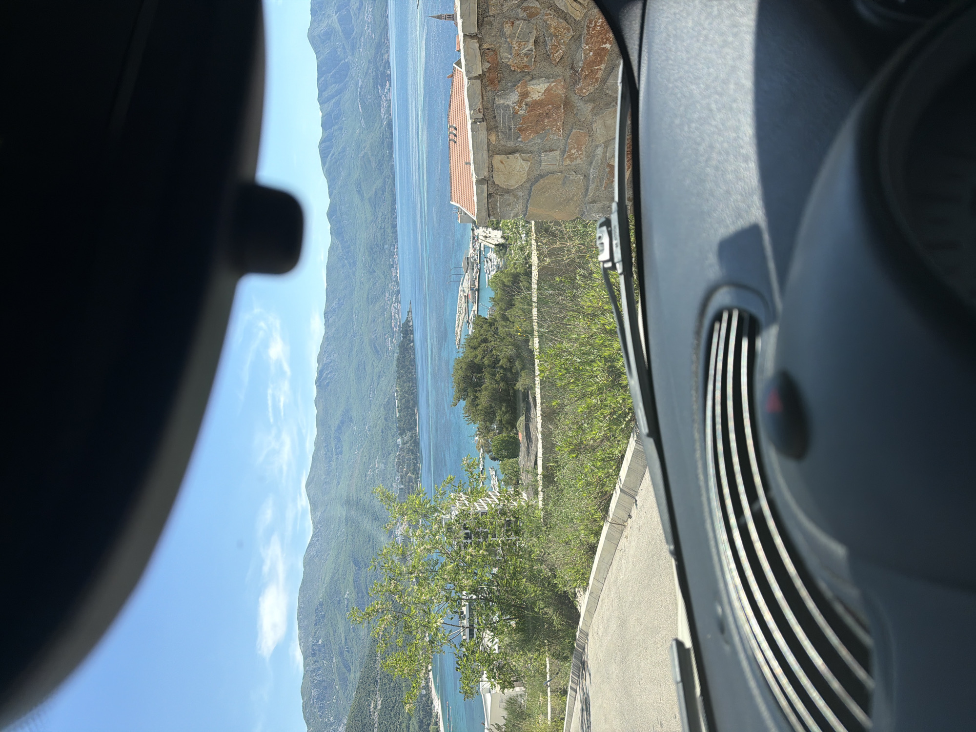 The image, taken from inside a car, shows a view of Budva, Montenegro. The car's interior, including the air vent and rearview mirror, frames the landscape outside. The Adriatic Sea stretches into the distance, bordered by mountains covered in green vegetation. A stone building with a terracotta roof is partially visible on the right. Below, a marina with several boats is visible. The sky is clear and blue. A tree is visible along the side of the road.