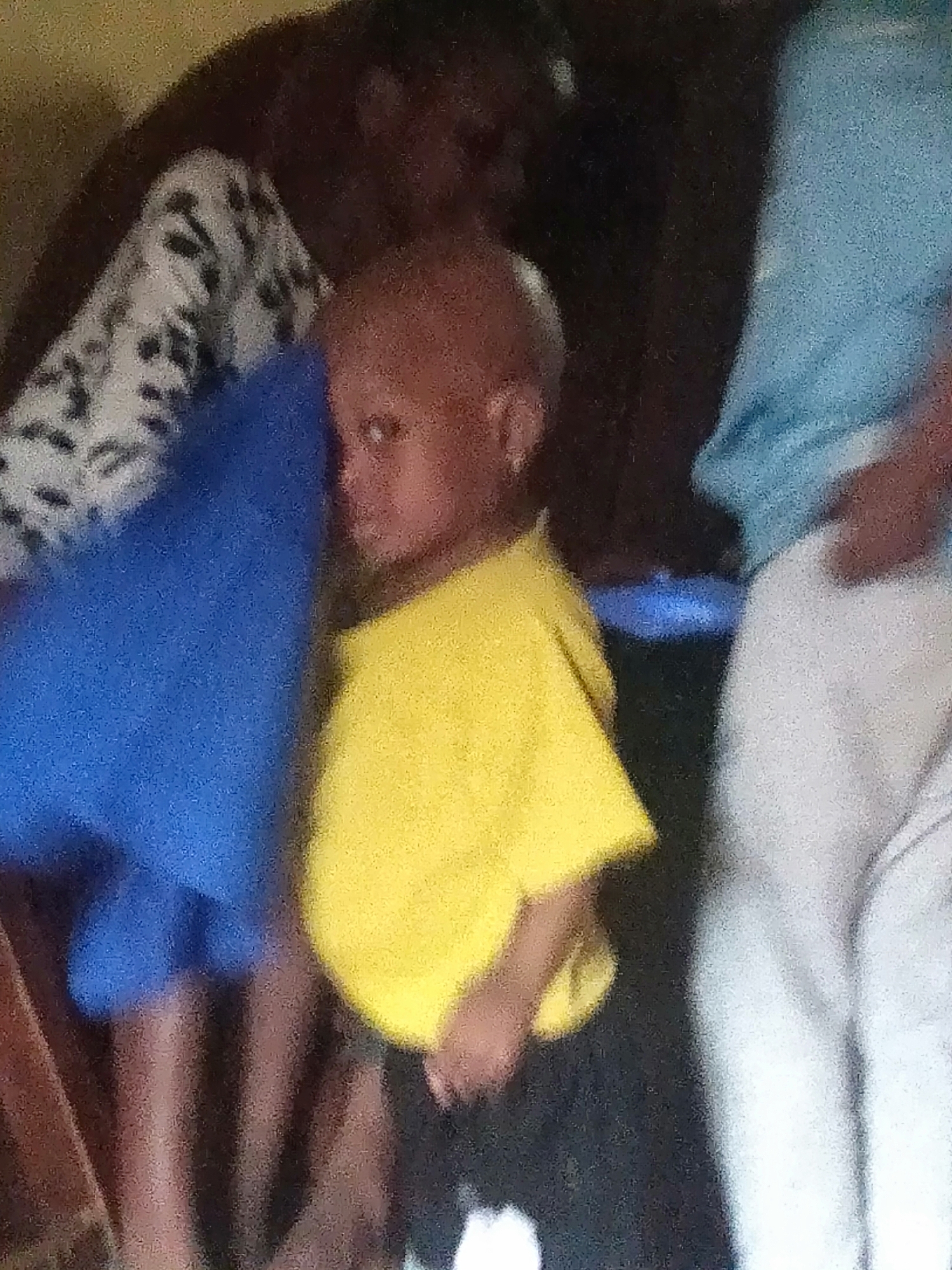 The grainy, low-resolution image captures an indoor scene with at least three individuals gathered together. In the center foreground stands a young child, possibly a toddler or preschooler, with short hair. They are wearing a bright yellow short-sleeved t-shirt and dark pants. The child's face is partially visible, turned slightly to the left with a neutral expression.
To the left of the child, another person is partially visible, wearing a white top with a black patterned design. A large, vibrant blue fabric object or piece of clothing is positioned in front of this person and extends towards the child. On the right side of the frame, another individual is seen from the torso down, wearing a light blue top and white pants.
The background is dark and indistinct, suggesting an indoor setting, possibly a room. A wooden-textured object, perhaps part of a chair or furniture, is faintly visible on the far left edge of the image. The subjects appear to be standing or seated in close proximity, indicating a casual gathering. The overall poor image quality, characterized by blurriness and visual noise, makes it difficult to discern specific details, time of day, or clear emotions. No text is visible in the image.