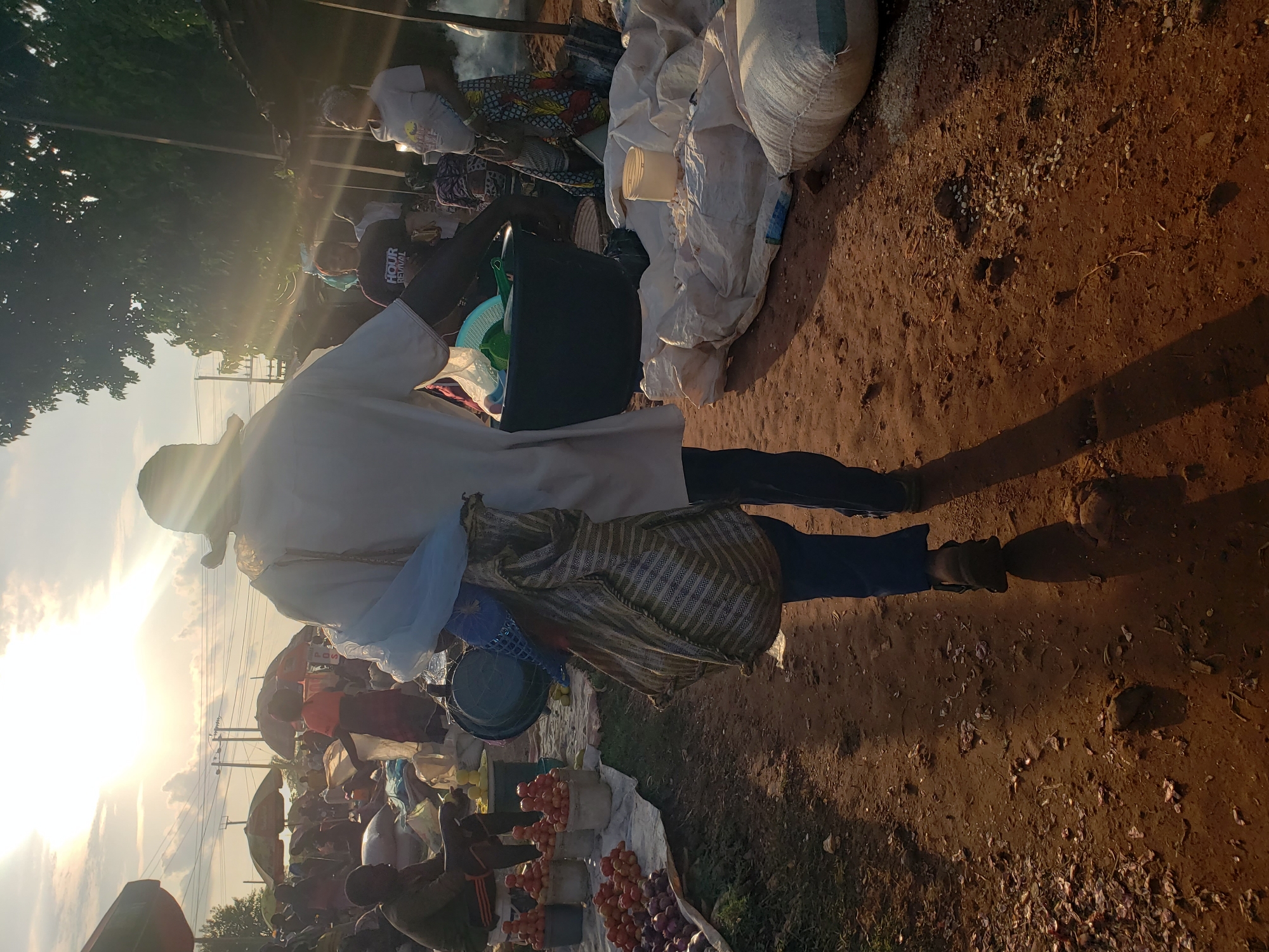 The image depicts a bustling outdoor market scene, likely in Kakuri Gwari, Nigeria, during the golden hour of sunset. The sun is low in the sky, casting long shadows and creating lens flare effects across the frame. In the foreground, a person walks away from the camera, carrying a large striped bag over their shoulder and a blue plastic bag in their hand. They are wearing a white shirt, dark pants, and a hat.
The market is filled with stalls and people. To the left, a vendor sits behind displays of red tomatoes piled in buckets and a large quantity of red onions. Other people are visible in the background, some standing and interacting, others tending to their wares. Various items are scattered around, including more buckets, sacks, and what appear to be food items. Umbrellas provide shade over some of the stalls. The ground is a mixture of dirt and scattered debris. The overall atmosphere is one of activity and commerce in a lively community setting.
