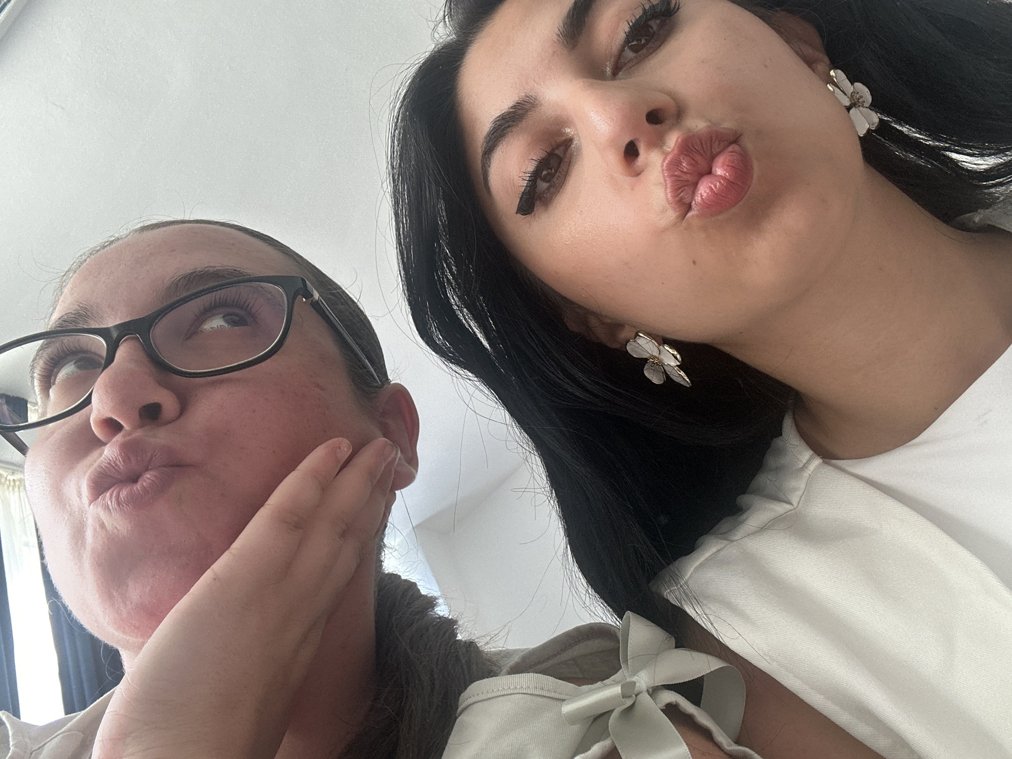 Two women are posing for a selfie. The woman on the right has long, dark hair and is wearing flower-shaped earrings and a white top with a bow. She has dark eyebrows, dark eyeliner, and is pursing her lips. The woman on the left is wearing glasses and has her hand on her cheek, also pursing her lips. The environment appears to be indoors. The image does not contain enough information to pinpoint a specific location in Plovdiv, Bulgaria.