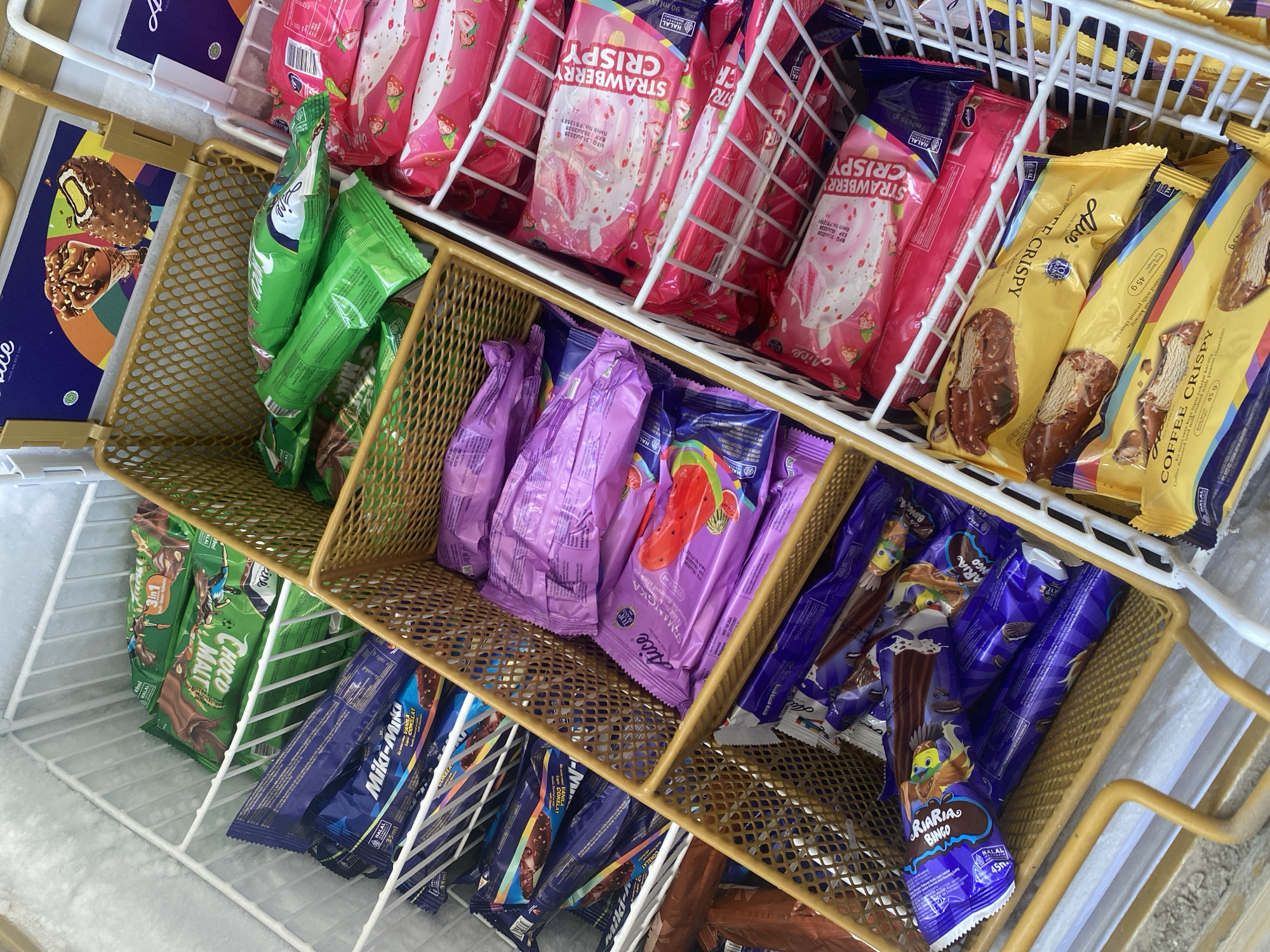 The image depicts an overhead view of an open freezer chest filled with various ice cream bars. The freezer is divided into compartments. In the top left, several green-wrapped ice cream bars labeled "Choco Malt" are visible. To their right, in a golden wire basket, are more green-wrapped ice cream bars. Below these, in a larger golden wire basket, are several purple-wrapped ice cream bars labeled "Aice Semangka" (watermelon flavor). To the right of the purple ice cream, in a white wire basket, are numerous pink-wrapped ice cream bars labeled "Strawberry Crispy." Below the purple ice cream, another golden wire basket holds blue-wrapped ice cream bars, some with cartoon bird imagery labeled "Riaria Bingo." To the right of this, in the white wire basket, are yellow-wrapped ice cream bars labeled "Coffee Crispy." The ice cream packaging suggests a retail environment, likely a convenience store or minimarket, possibly located in Bantaeng, Indonesia, given the Halal certification visible on several packages and the common availability of these brands in Southeast Asia. The freezer is frosted, indicating it is a functional refrigeration unit. There are no people or discernible actions occurring in the image.