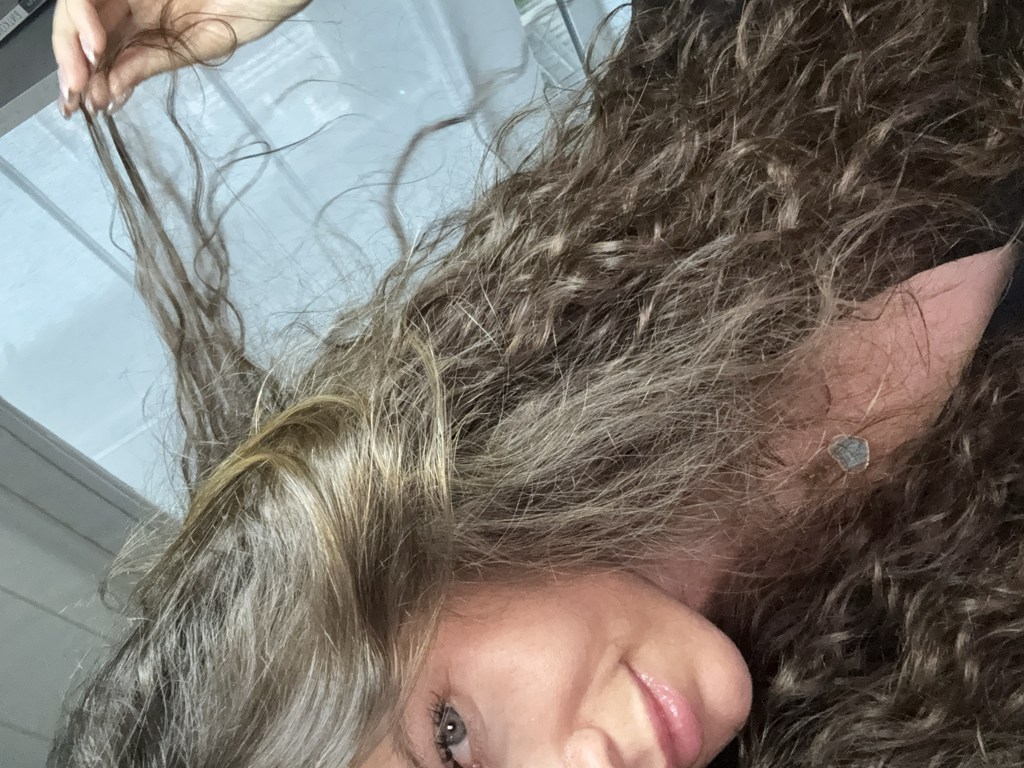 The image is a close-up, low-angle, horizontal shot of a woman's face and her long, dark, curly hair. She is partially visible, with her left eye and lips in focus. Her eye is blue-grey and she is smiling subtly. Her lips are glossy pink. Her hair is dark brown with lighter brown and blonde highlights. Her right hand is visible, holding a few strands of her hair near the top of her head. The background appears to be a window with vertical blinds or a similar structure, suggesting an indoor setting, potentially a home or an office. There is no discernible information to place this image in Moscow, Russia.