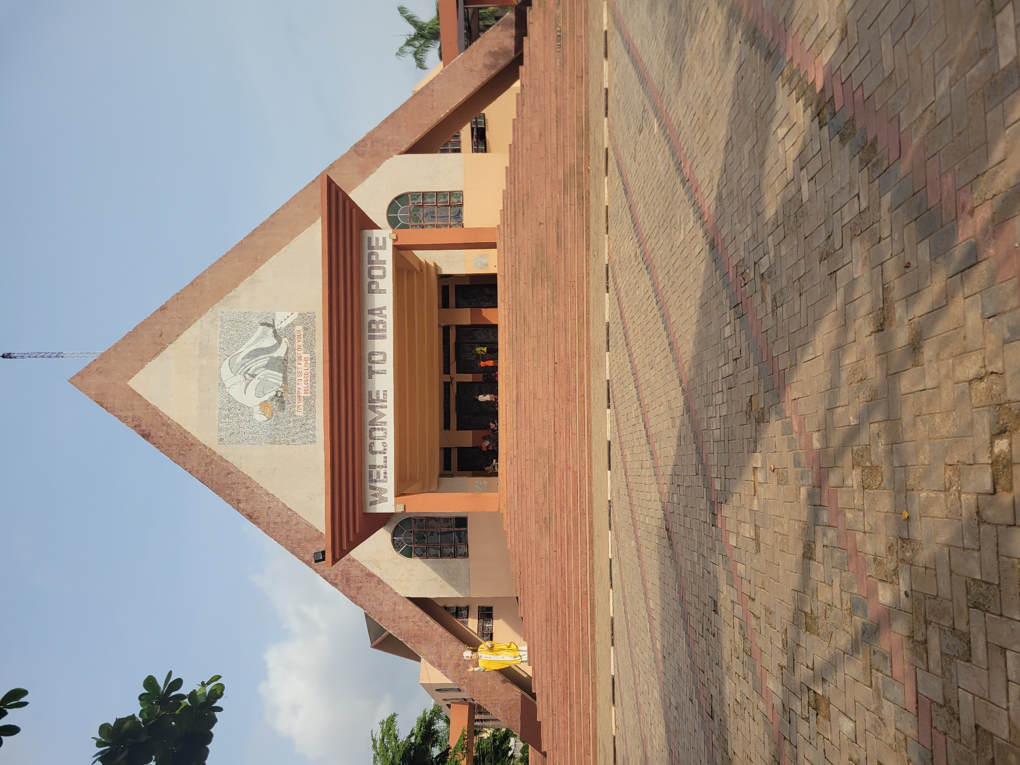 The image depicts the entrance of the Pope John Paul II Major Seminary in Onitsha, Nigeria. The building features a large triangular facade with a mosaic of Pope John Paul II and the inscription "WELCOME TO IBA POPE." There are stained-glass windows on either side of the entrance. A statue of a figure is visible to the left. The foreground is composed of a patterned brick walkway leading up to the building. The sky is partially visible with some trees and clouds.