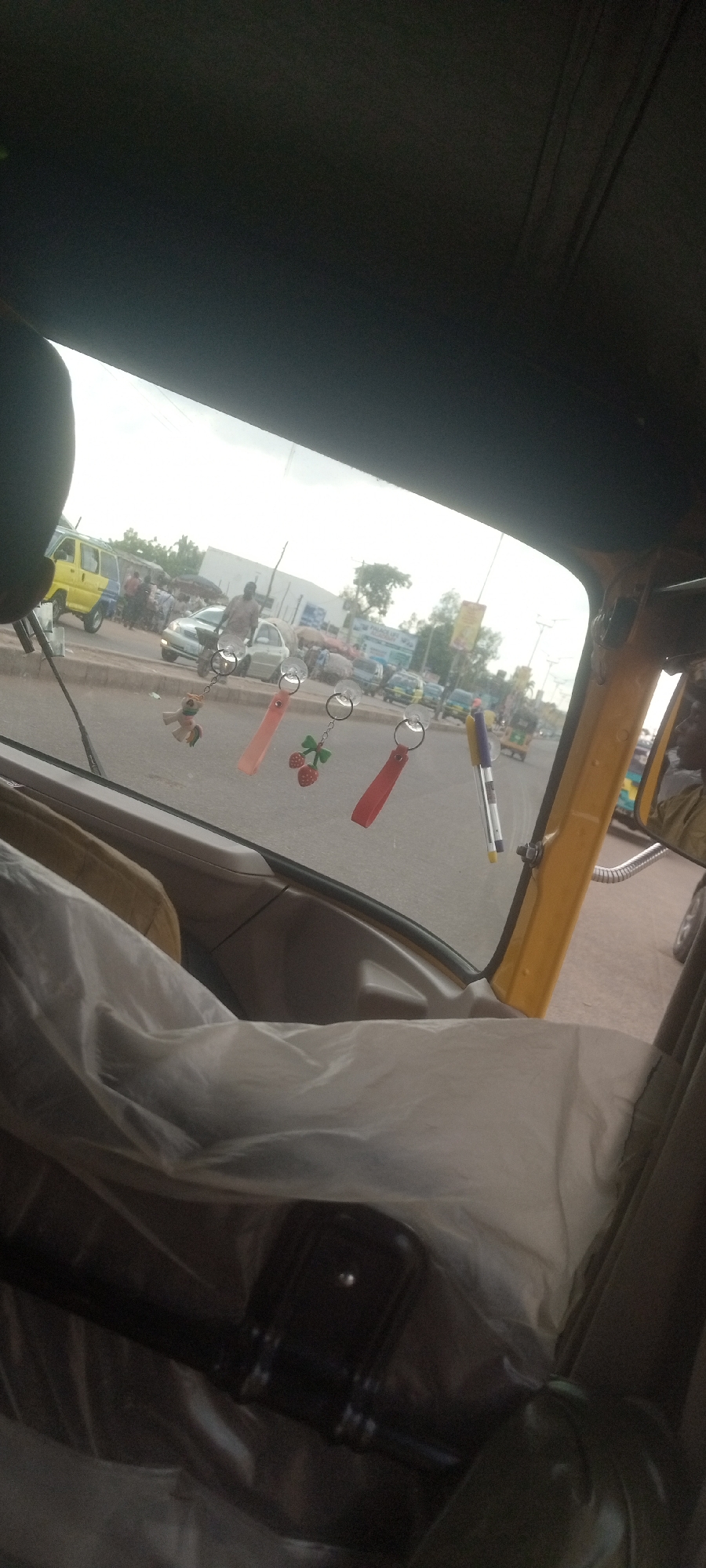The image shows a view through the windshield of a vehicle, possibly an auto-rickshaw (keke), in Sabon Tasha, Nigeria. Hanging from the windshield are four items attached with suction cups: a small horse or pony toy, two rectangular keychains, and a pen. The seats in the vehicle are covered in a translucent, possibly plastic, material. Outside, the street is busy with people and vehicles, including a yellow van and a number of cars. A building can be seen in the background, and a billboard or sign is visible on the right side of the street. There are people visible on the sidewalks on both sides of the road.