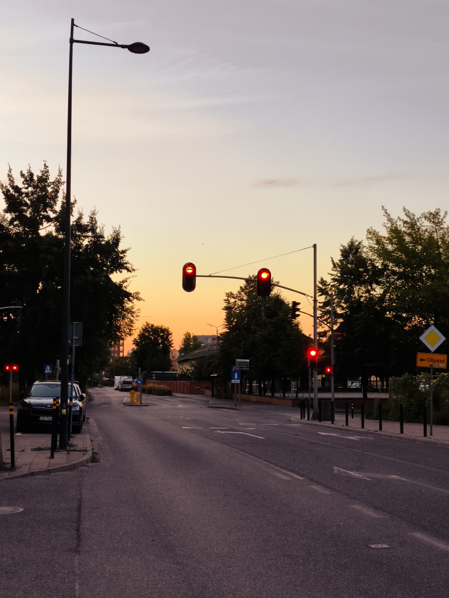 The image depicts a street scene at dusk in Gdańsk, Poland, likely near the city center given the presence of traffic lights and urban vegetation. A dark-colored SUV is parked on the right side of the street, its license plate reading "GD 7AA". Multiple traffic lights are illuminated red, indicating a stop signal. Tall street lamps and mature trees line the asphalt road, which has visible lane markings. In the background, buildings and more trees are visible under a sky transitioning from twilight hues to a pale, warm orange. A yellow diamond-shaped sign with a black arrow and the Polish word "Objazd" (detour) is posted on the right side of the road.