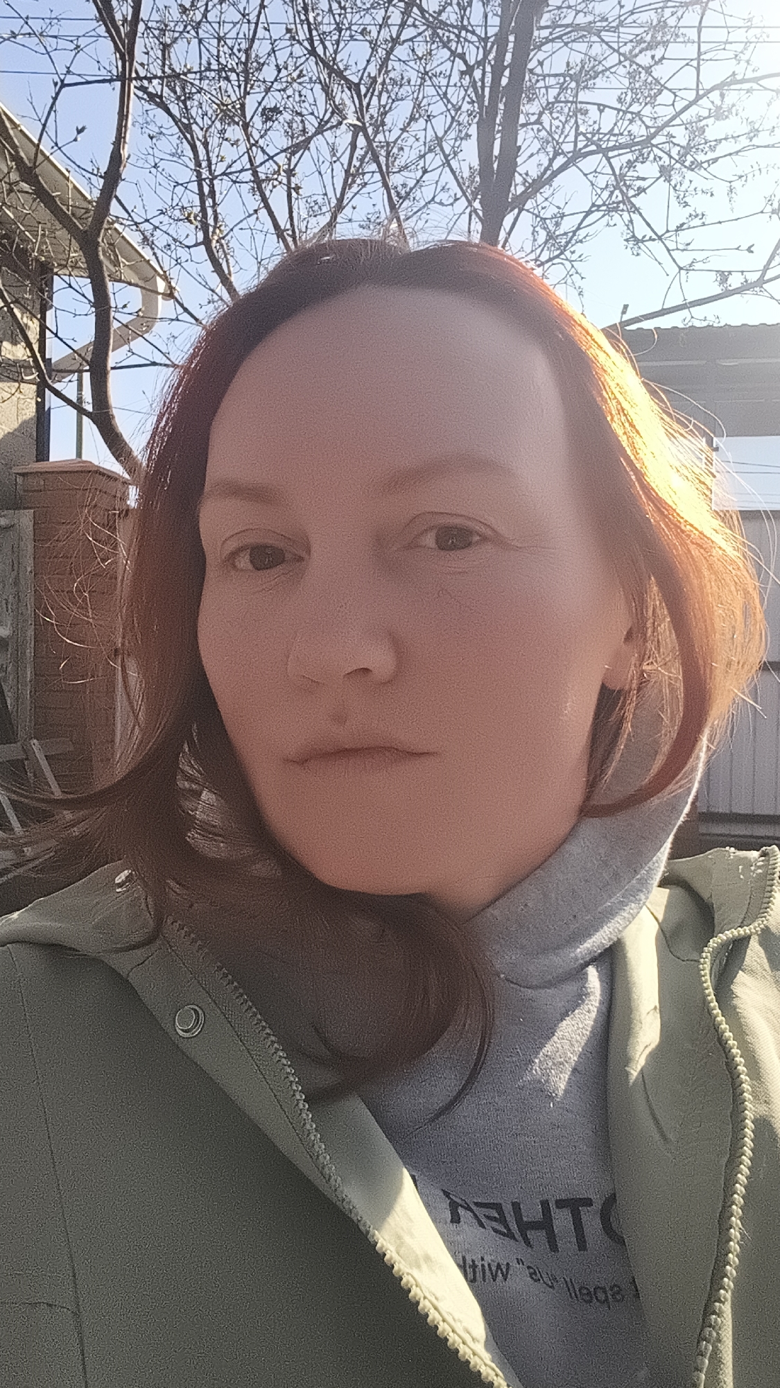 A close-up selfie captures a woman outdoors in Chaikovsky, Russia, during what appears to be a sunny day, likely in late winter or early spring given the bare tree branches. The woman, with reddish-brown hair, looks directly at the camera with a neutral expression. Her hair is brightly lit by sunlight on the right side of the frame, suggesting a direct light source. She is wearing a light green or sage green jacket over a grey hooded sweatshirt. Partially visible and reversed text on her grey sweatshirt can be discerned as "OTHEA" and "JiW 's' lleps" (likely "OTHER" and "spell 's' with J" or similar). The background consists of intricate bare tree branches silhouetted against a clear blue sky. Parts of a building structure and a fence or gate are also visible, indicating an outdoor, possibly residential, setting. The strong sunlight creates clear highlights and suggests the time of day could be morning or late afternoon.