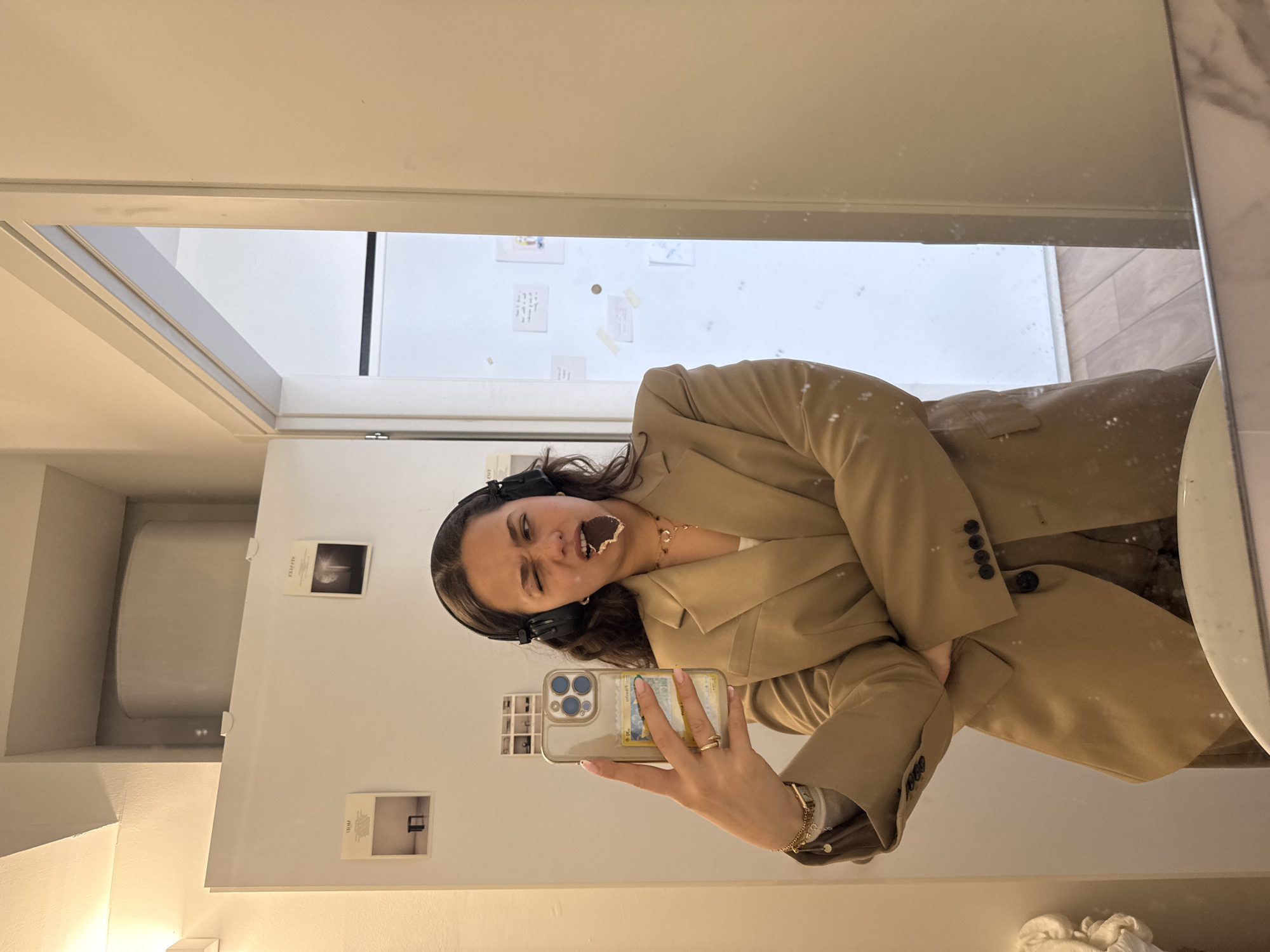 This mirror selfie, taken in Milan, Italy, captures a young woman making a playful expression. She has long dark hair and is wearing a beige blazer and black over-ear headphones. A piece of dark chocolate or snack is partly in her mouth, with some residue on her cheek, and she is giving a slight squint or wink. She holds a smartphone, likely an iPhone, encased in a clear cover that prominently displays a Pokémon card, identifiable by "Pikachu" and Japanese text. She also wears a gold ring and a gold bracelet.
The background, visible through the mirror, features white walls adorned with several small, square photos or notes. One photo appears to show a bright light or moon, while another depicts a dark room with a single chair. On the left side of the reflection, a large white cylindrical object is partially visible. A strip of marble-patterned trim frames the right side of the mirror. The indoor setting suggests a casual, personal moment.