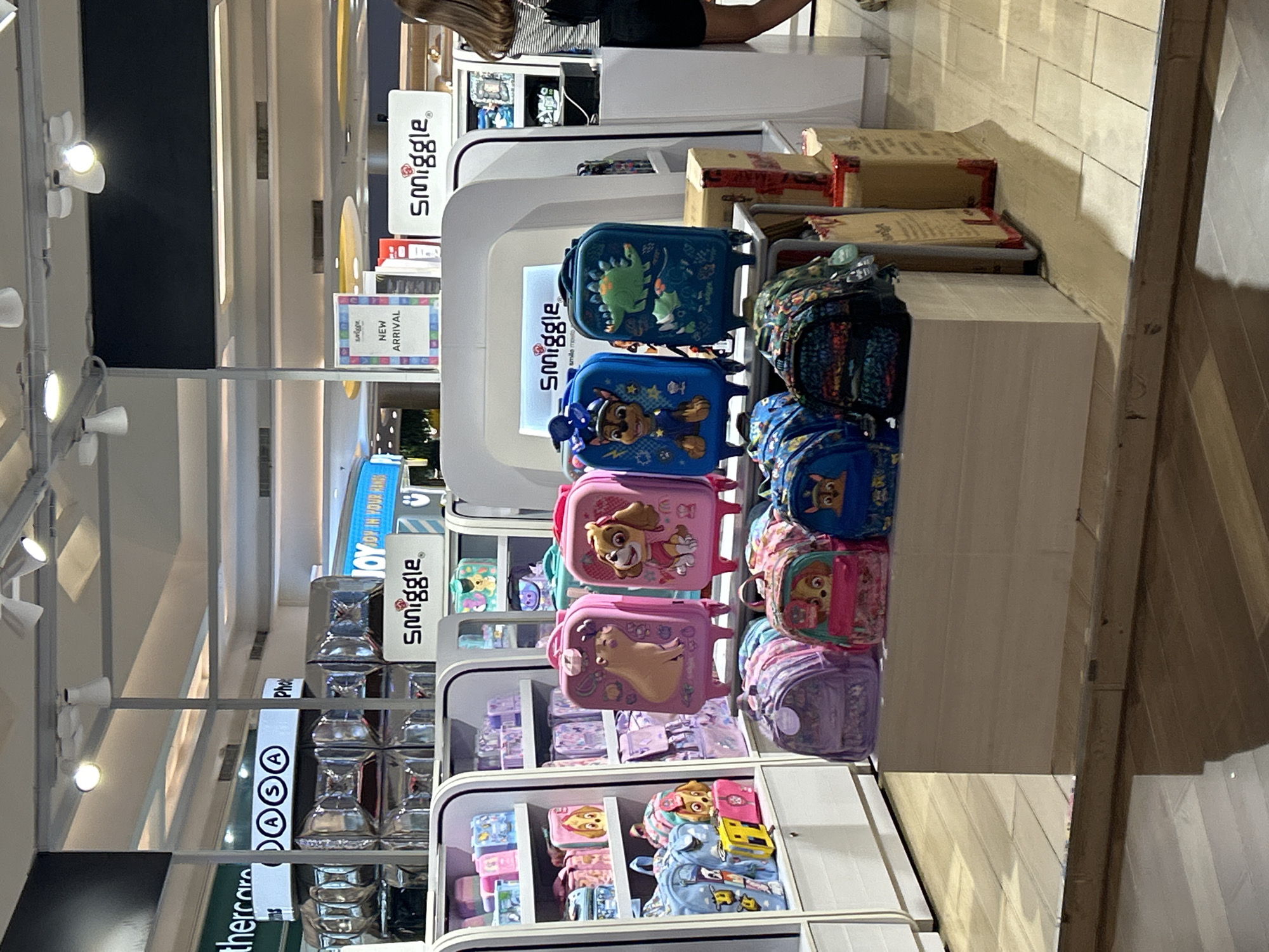 The image depicts the interior of a retail store, likely a Smiggle branch, featuring an array of children's backpacks and wheeled suitcases. The merchandise is displayed on shelves and stands within a brightly lit store environment. The flooring appears to be tiled. Signs for "Smiggle" are visible throughout the store. A partially visible woman with long brown hair, wearing a striped shirt and dark shorts, is standing to the right of the frame, facing away from the camera. Based on the presence of the Smiggle store, this location is likely within a mall in Makassar, Indonesia.