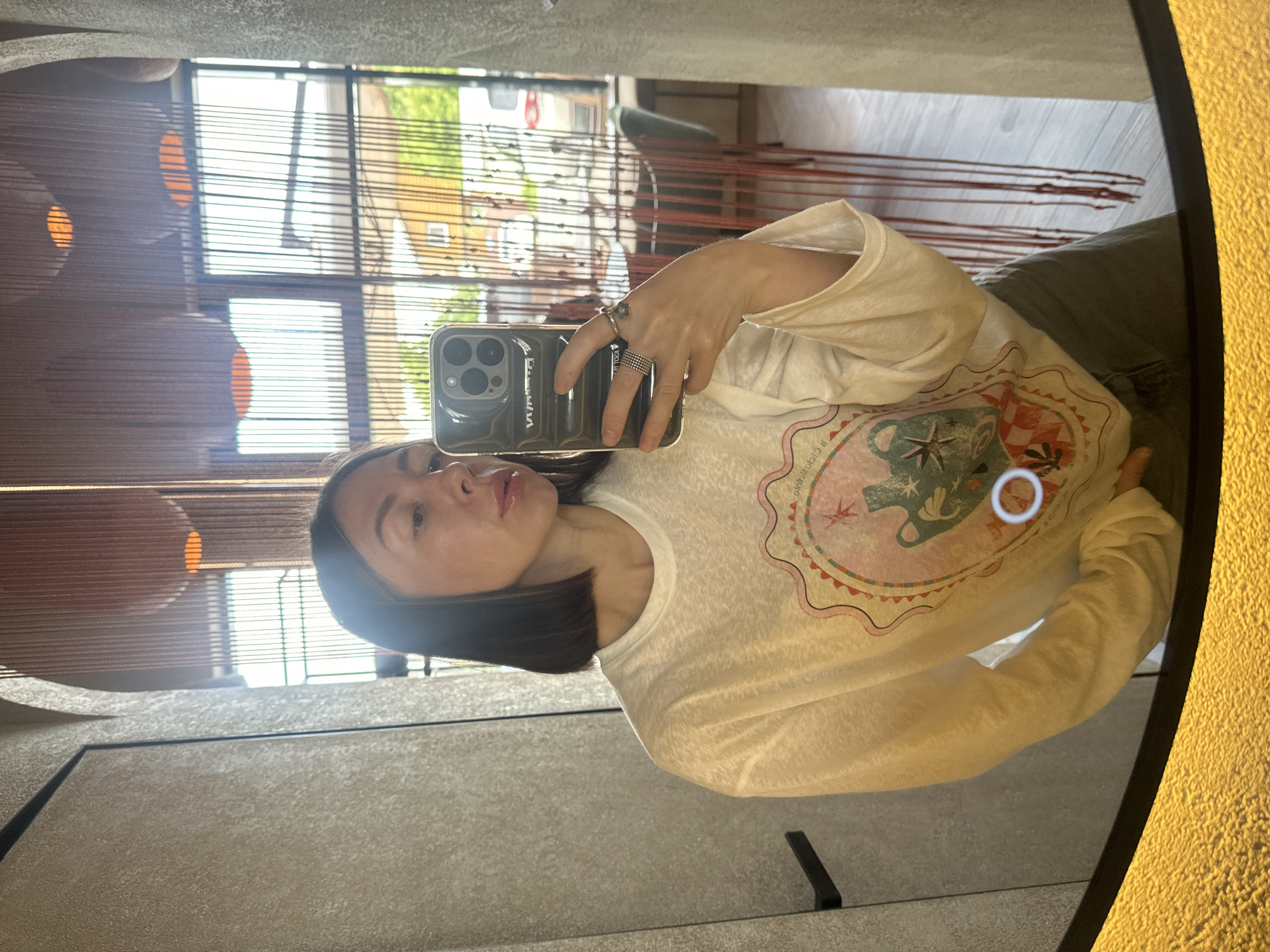 A medium shot captures a woman taking a mirror selfie in an indoor setting in Усады, Russia. The woman, with short, straight dark hair, is reflected looking towards the camera, holding a dark-cased smartphone (likely an iPhone) with her right hand, which displays multiple rings. She wears a white, slightly translucent, long-sleeved t-shirt featuring a colorful graphic design on the front depicting a framed image with an amphora and a star. Dark-colored pants are partially visible below her shirt.
The mirror's reflection reveals an interior with a textured light-colored wall and a dark vertical trim to the left, and an archway above. In the background, beyond the woman, large windows are visible, partially obscured by a brown, vertical string-like curtain or decorative divider. Through the windows, an outdoor scene is reflected, showing green foliage, residential buildings, and a white van. Interior elements reflected include recessed circular lights and what appears to be dining furniture (chairs and tables) in the distance. The bottom edge of the mirror features a distinctive textured, golden-yellow border. A white circular highlight is visible on the graphic design of the woman's shirt.