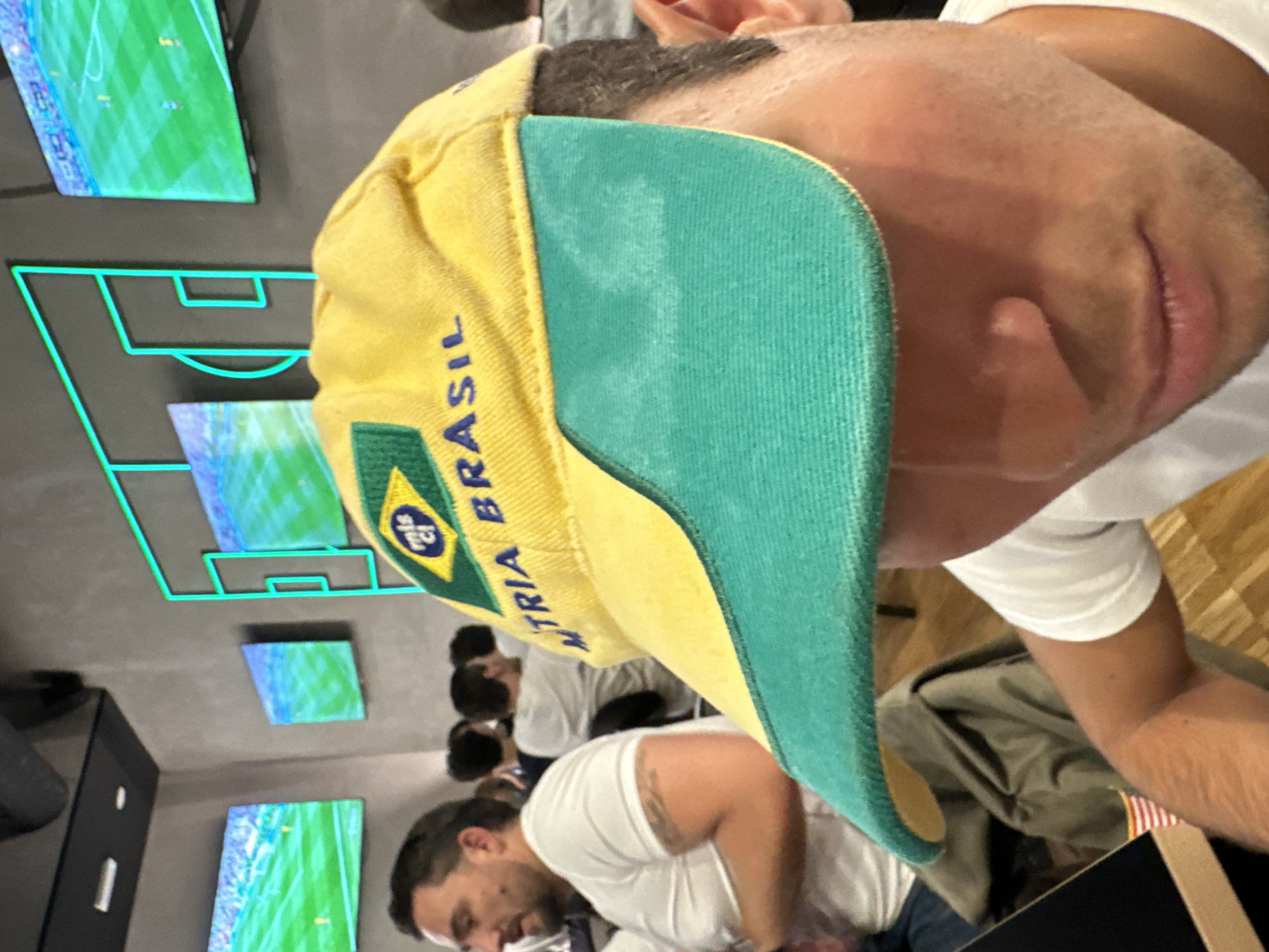 A man wearing a yellow and green Brazil-themed cap is the primary subject, with the cap obscuring his eyes and forehead. The cap has the word "BRASIL" written on it in blue letters and a small circular emblem with "mls cl" inside. The man is wearing a white t-shirt. In the background, several other people are visible, also appearing to wear white t-shirts. They are seated, suggesting a public space like a restaurant or bar. Mounted on the wall behind them are multiple flat-screen televisions displaying what appear to be soccer matches, indicated by green fields and players. The wall also features a stylized graphic resembling a soccer field in neon blue light. The environment appears to be indoors, with a muted color palette and wooden flooring visible. The overall scene suggests patrons watching a sporting event, likely soccer, in a commercial establishment. The presence of the neon soccer graphic and multiple screens could indicate a sports bar.
