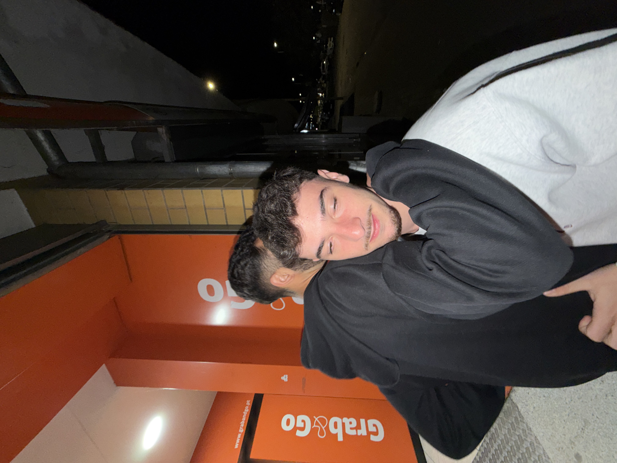 Two young men are embracing in front of a "Grab&Go" vending machine, likely in Bragança, Portugal. One man, with dark hair, is wearing a black hoodie and black pants. The other man, also with dark hair, is resting his head on his friend's shoulder and is wearing a grey and black hoodie. They are standing on a paved surface in an outdoor environment, possibly at night, given the darkness and street lights visible in the background. A building with orange panels and the "Grab&Go" vending machine are visible behind them.