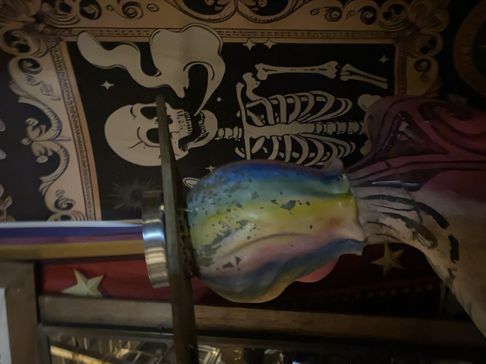 The image shows a close-up of a shelf or display, likely indoors. The backdrop features a black and white drawing of a skeleton exhaling smoke, framed by ornate designs. In front of the backdrop is a multi-colored, possibly ceramic, object resembling a head, positioned behind a vertical metal pole. Below the head, there is a red surface with a gold star. The location is Baan Nai Suan, Thailand.