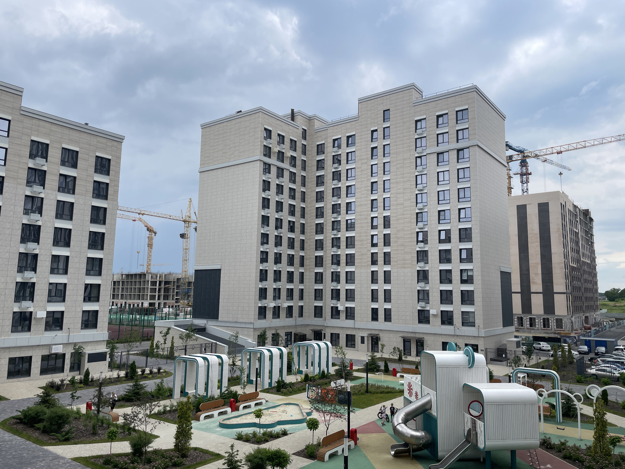 This media file captures an outdoor scene in a residential complex, likely during the daytime with overcast weather. In the foreground, a modern playground features colorful surfaces, a sandpit, benches, and abstract play structures with slides. Two children are visible playing on the playground, one near a slide and another on a bicycle.
The scene is framed by several multi-story apartment buildings with light-colored facades and numerous dark-paneled windows. In the background, construction cranes are active, indicating ongoing development in the area. The architectural style of the buildings appears modern and uniform. The presence of parked cars and a road suggests an urban or suburban setting.
The overall impression is one of a developing community with a focus on modern amenities and family living. The cloudy sky suggests a cool or mild day, with no distinct shadows indicating a specific time of day. The location is identified as Stavropol, Russia. No visible text is present in the image.