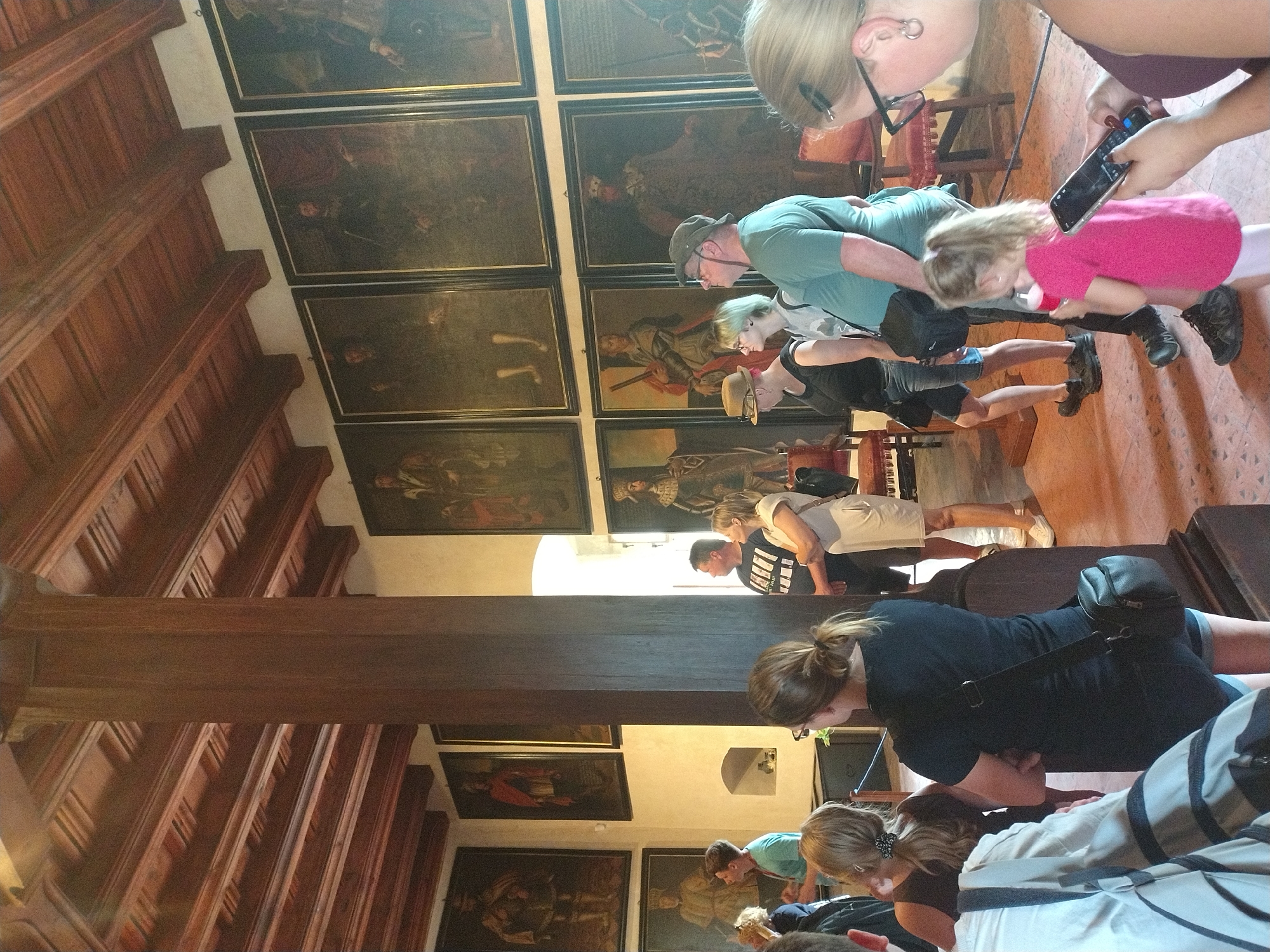 The image depicts a group of tourists inside a room with a high, wooden-beamed ceiling and ornate paneling, likely a historical building. Several large, framed paintings of historical figures in formal attire adorn the walls. In the foreground, a man wearing a green t-shirt and a cap is looking up at a painting, while a woman with blonde hair in a black t-shirt stands beside him. A young girl in a pink shirt is visible in front of them, holding a red cup. To the left, a man in a blue shirt is speaking to a group of people, and a woman with blonde hair in a black top is observing. The room is furnished with chairs and appears to be a museum or gallery in Karlštejn, Czechia. The lighting suggests it is daytime.